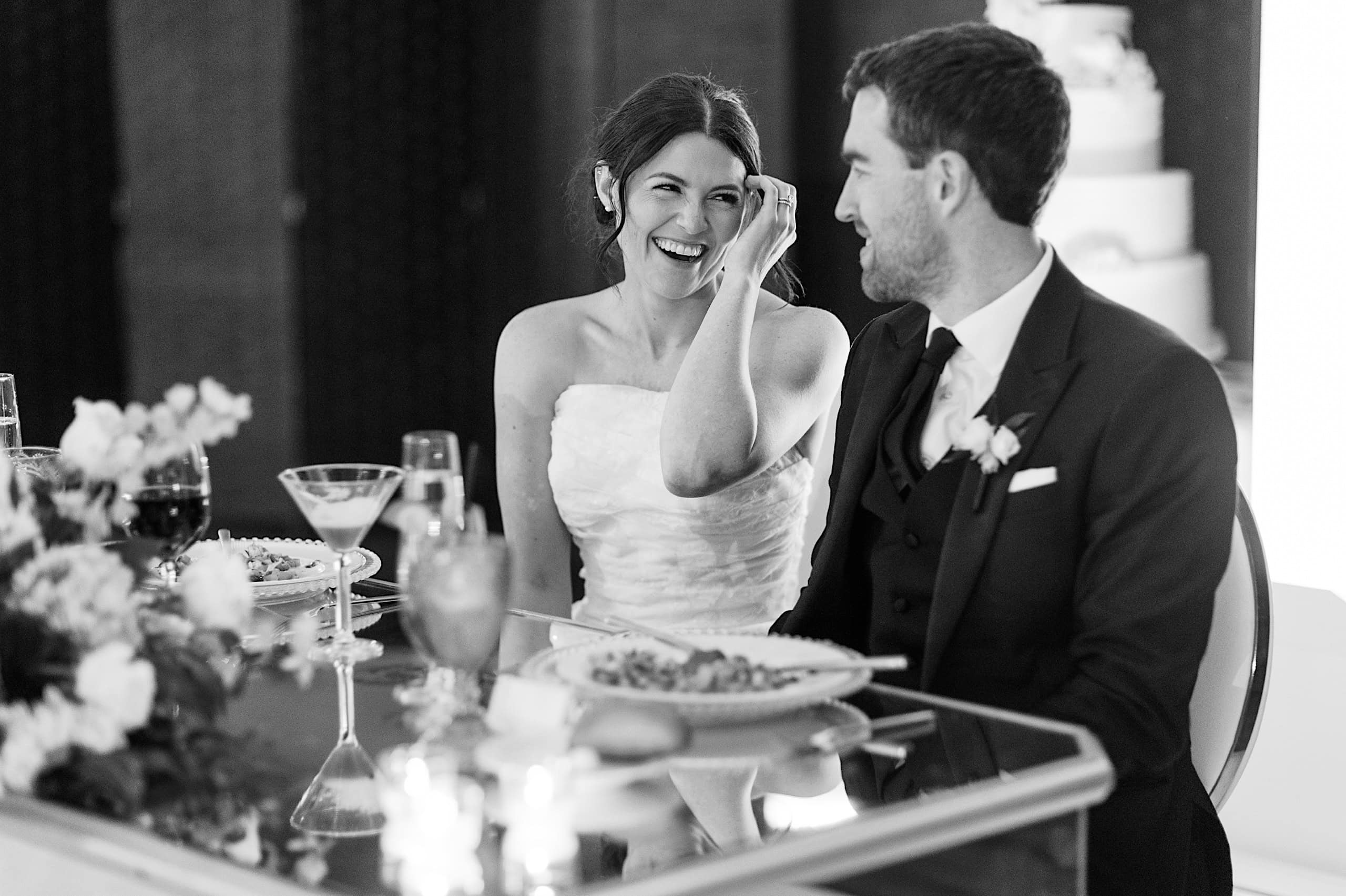 A bride and groom sit at a table, smiling and talking during their elegant spring wedding reception at The Ivy at Ellis Preserve. Plates of food and drinks are in front of them, capturing a joyful moment.