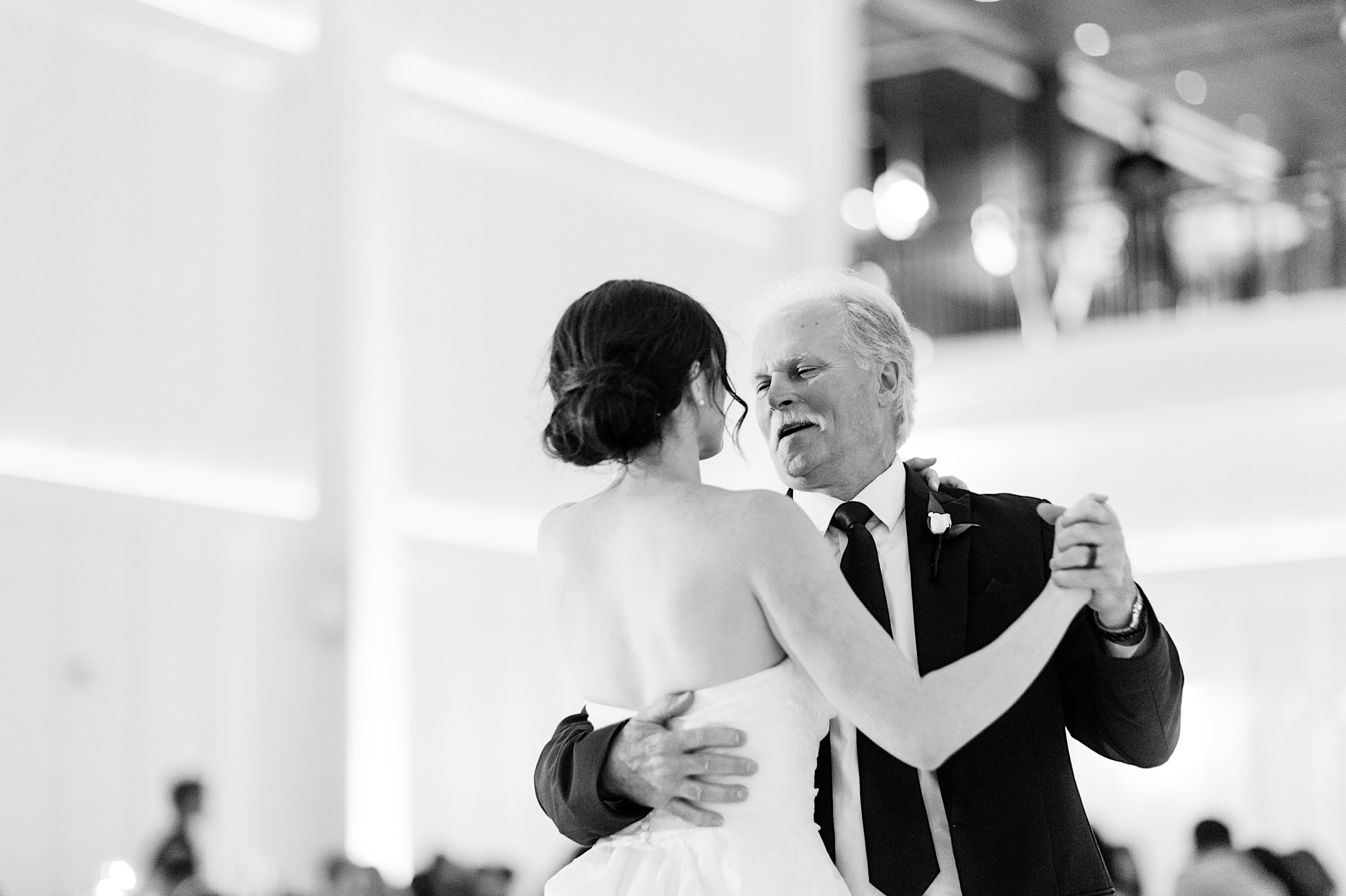 At The Ivy at Ellis Preserve spring wedding, a bride in a strapless dress dances with an older man in a suit at a formal indoor event, both smiling as they gaze at each other.