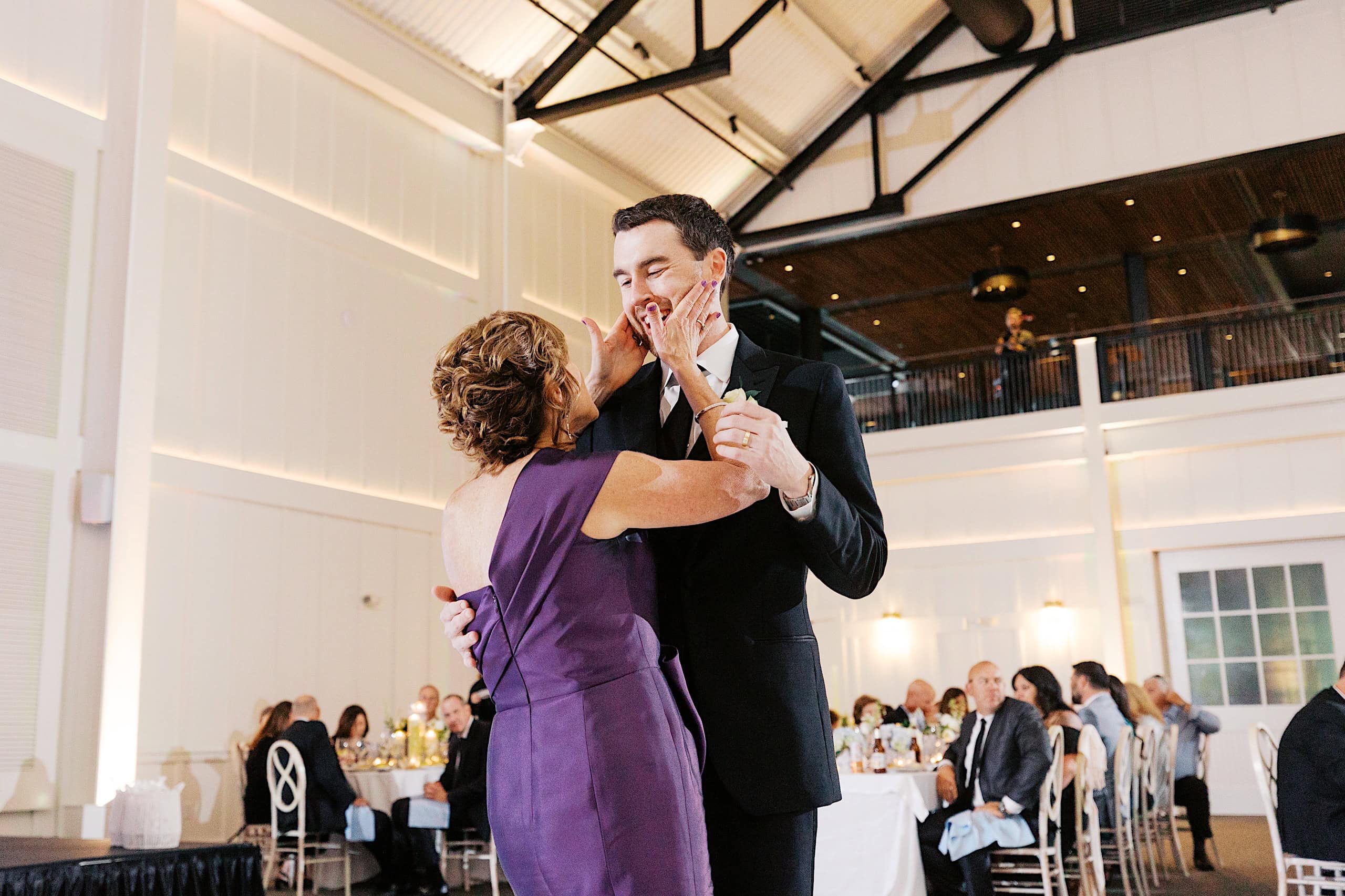 A woman in a purple dress and a man in a suit share a dance at The Ivy at Ellis Preserve spring wedding, surrounded by seated guests in a bright, modern venue.