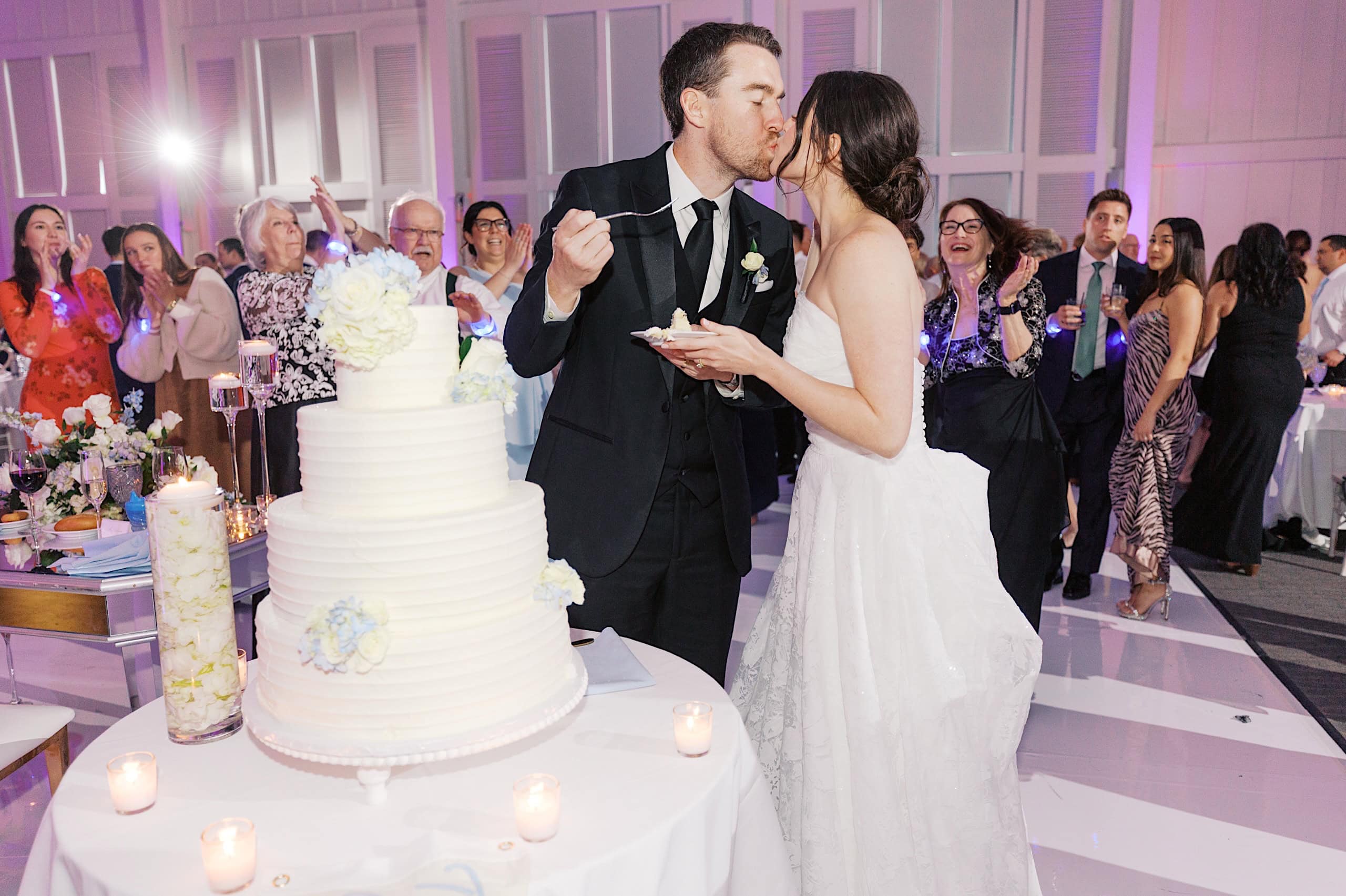Bride and groom kiss while feeding each other cake at their The Ivy at Ellis Preserve spring wedding reception, surrounded by guests. A large tiered cake with flowers is displayed in the foreground.