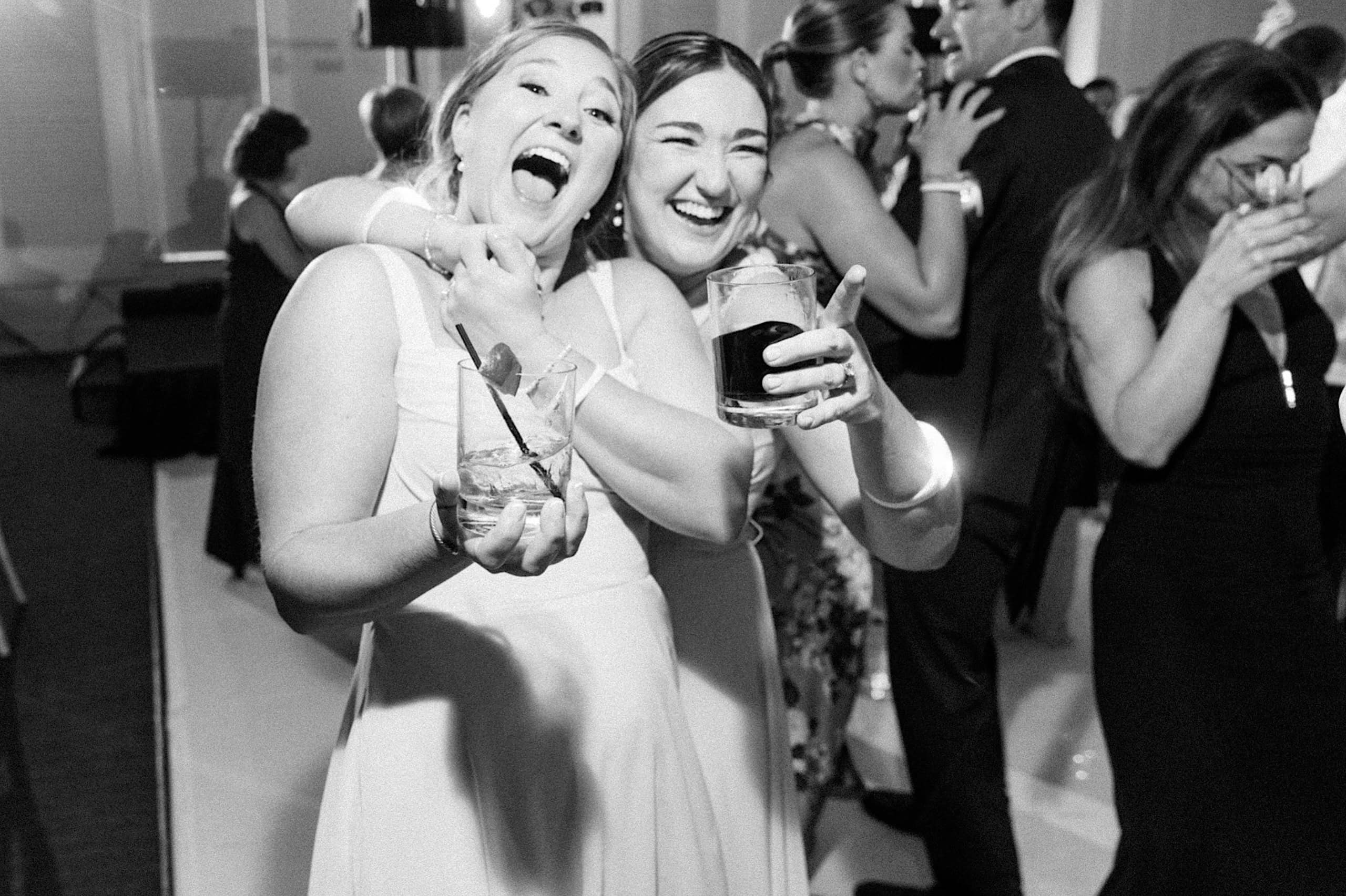 Two women in dresses smile and hold drinks at a lively indoor event during The Ivy at Ellis Preserve spring wedding, surrounded by guests who are dancing and celebrating.