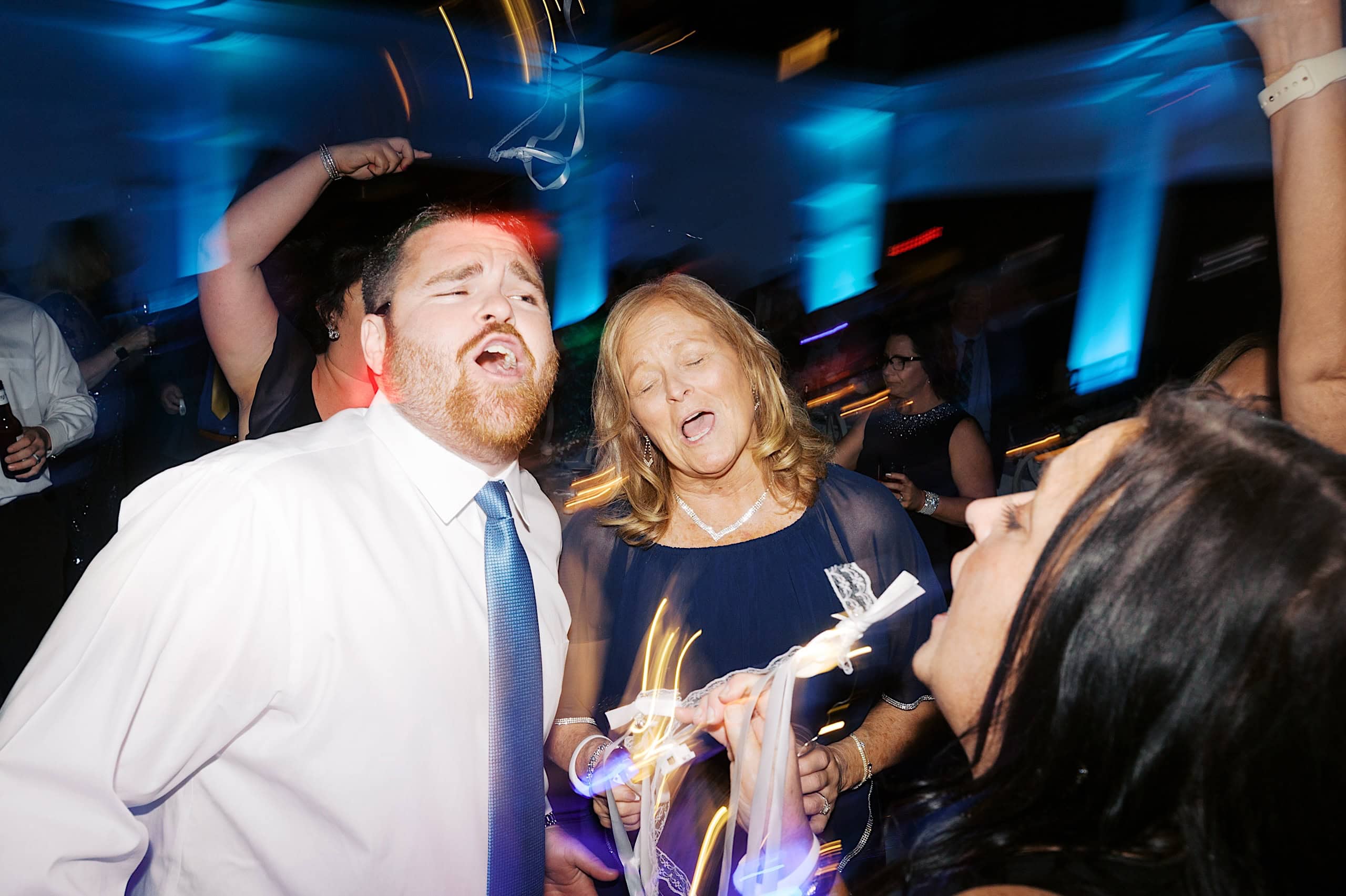 A group of people in formal attire sing and dance energetically at a party with blue lighting and light streaks, capturing the lively spirit of The Ivy at Ellis Preserve spring wedding celebration.