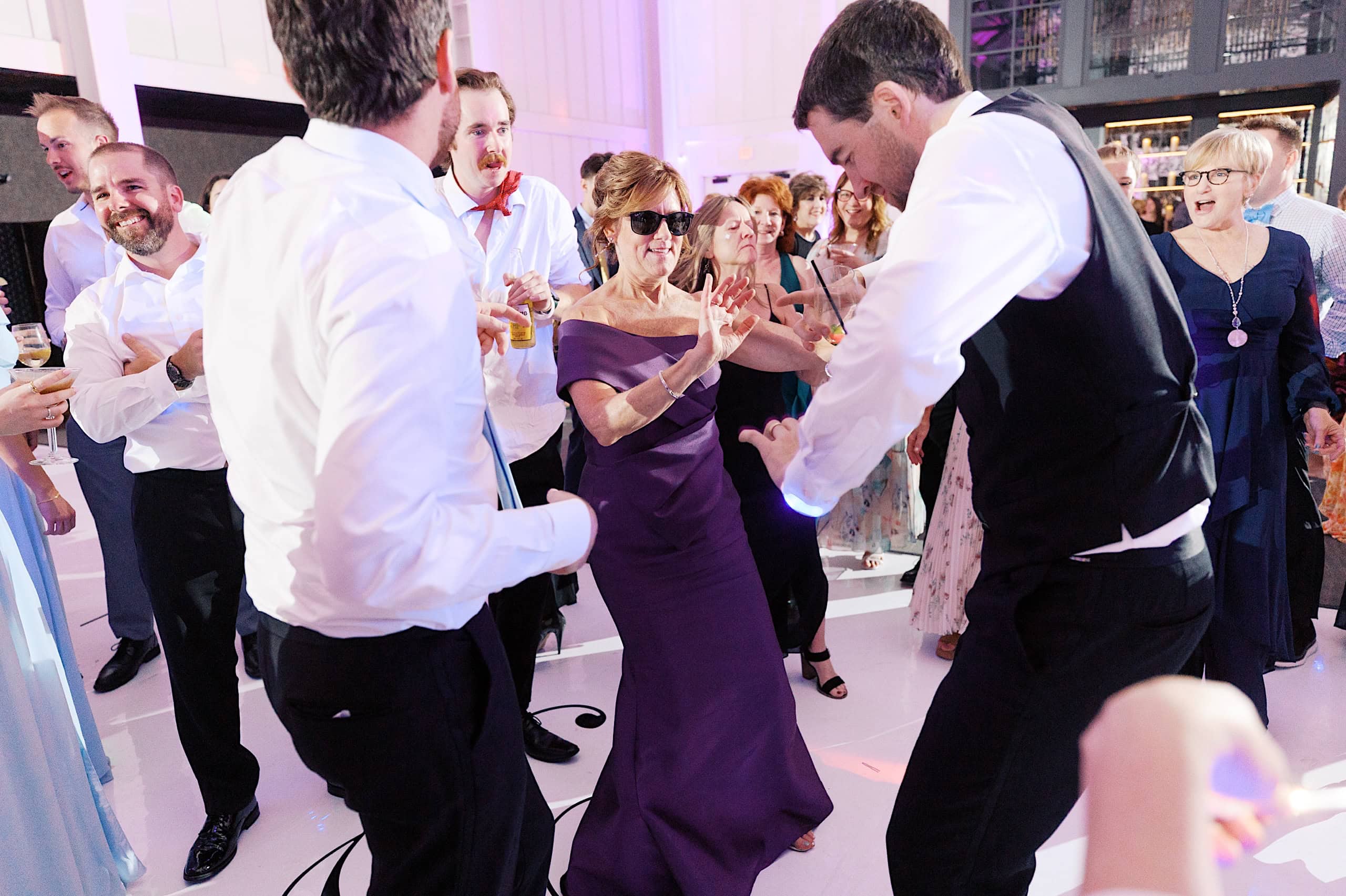 A group of people, dressed in formal attire, dance and celebrate together indoors under bright lighting at The Ivy at Ellis Preserve spring wedding.