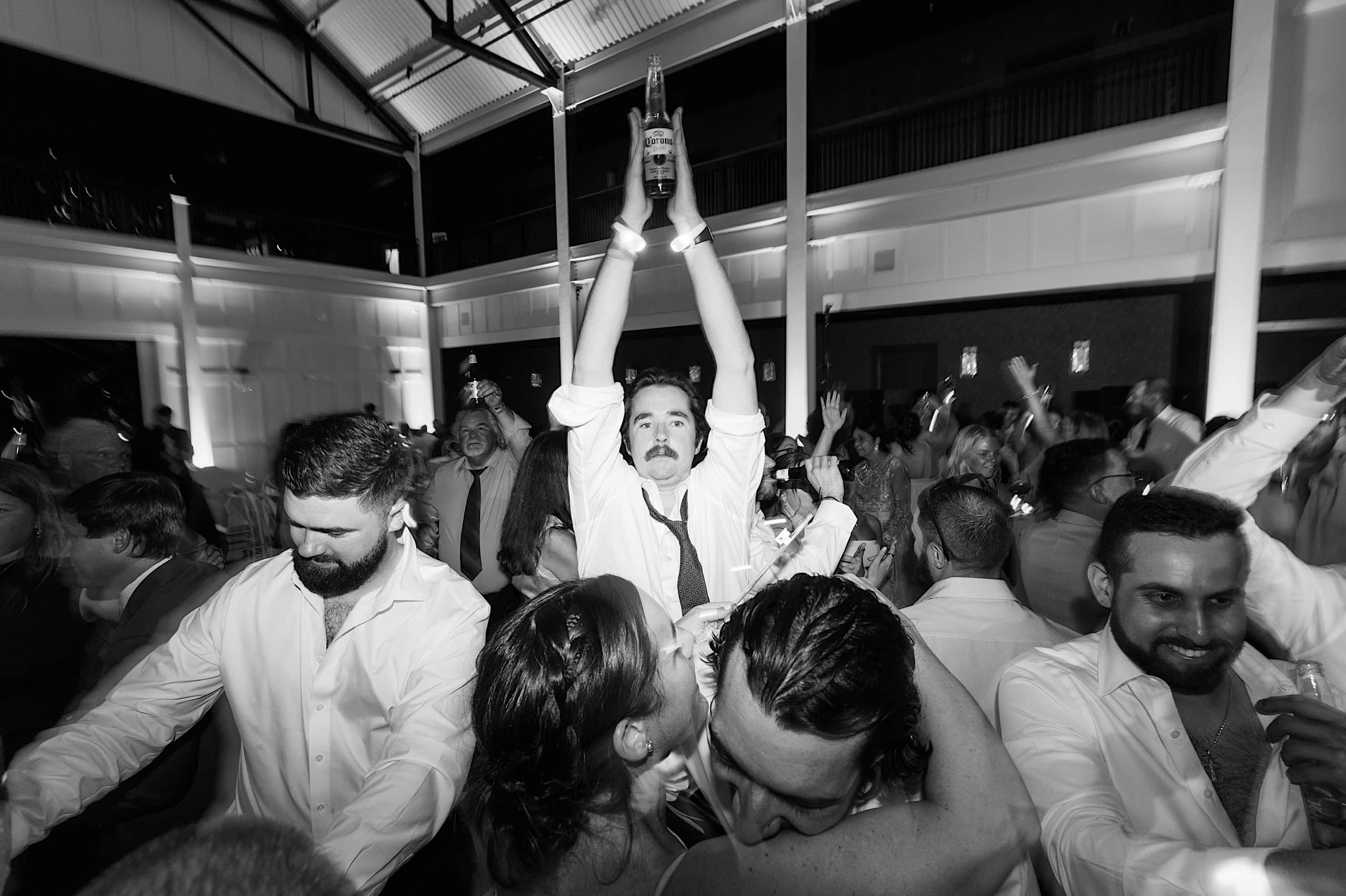 At a lively indoor party, reminiscent of The Ivy at Ellis Preserve spring wedding celebrations, a man in a dress shirt raises a bottle above his head as people around him dance and celebrate.