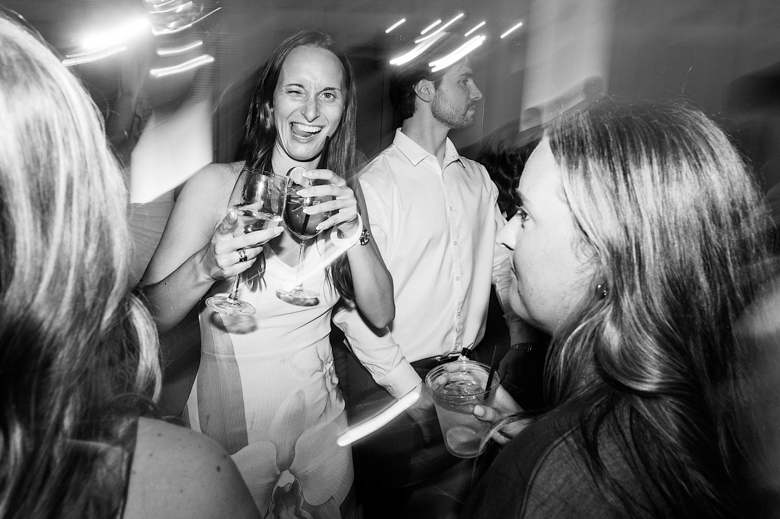 At a lively The Ivy at Ellis Preserve spring wedding, a woman in a white dress holding drinks smiles and winks at a party, surrounded by others. The black and white image features stunning motion blur effects.