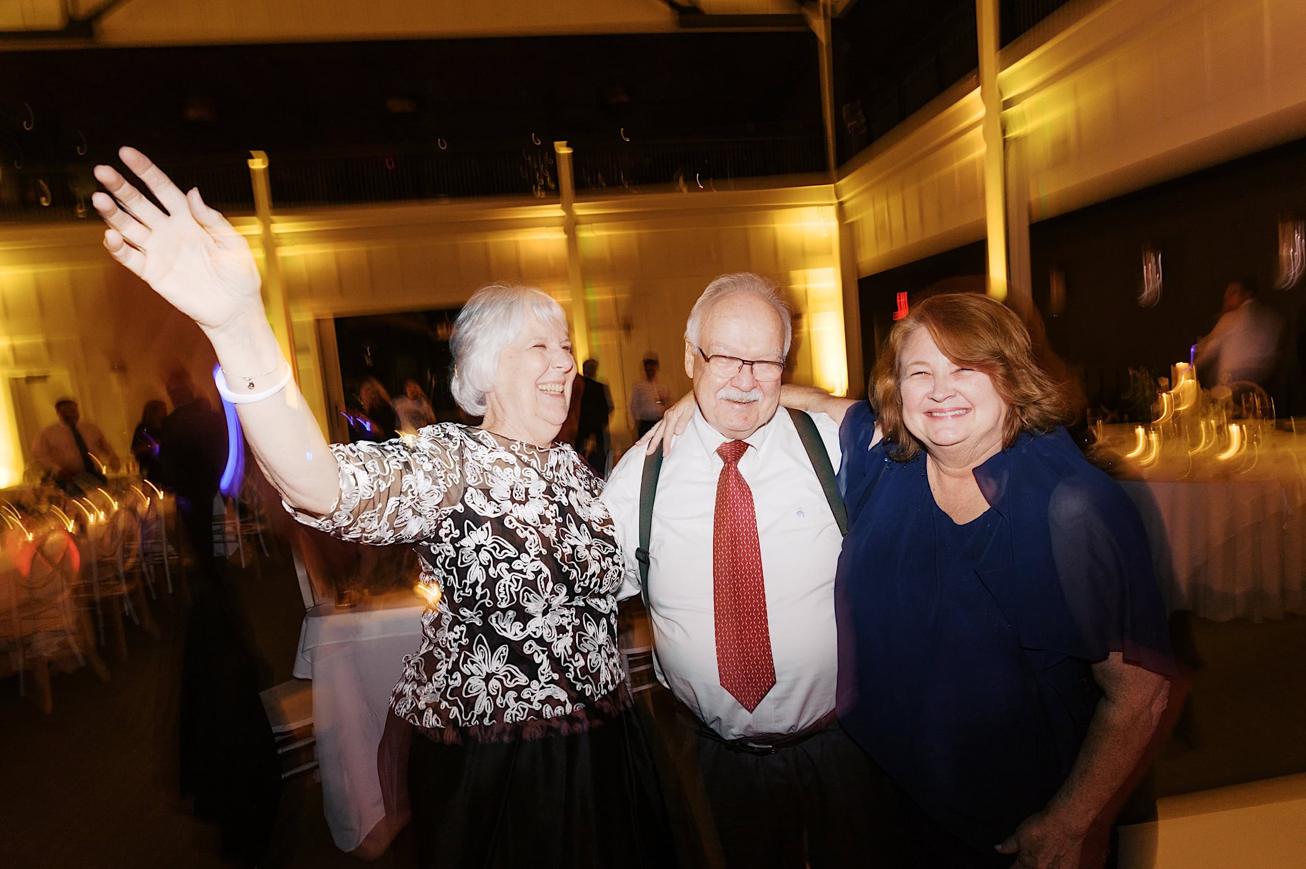 Three older adults stand together indoors at an event, smiling; one woman raises her arm in celebration. The background glows with warm lighting and tables, capturing the joy of a The Ivy at Ellis Preserve spring wedding.
