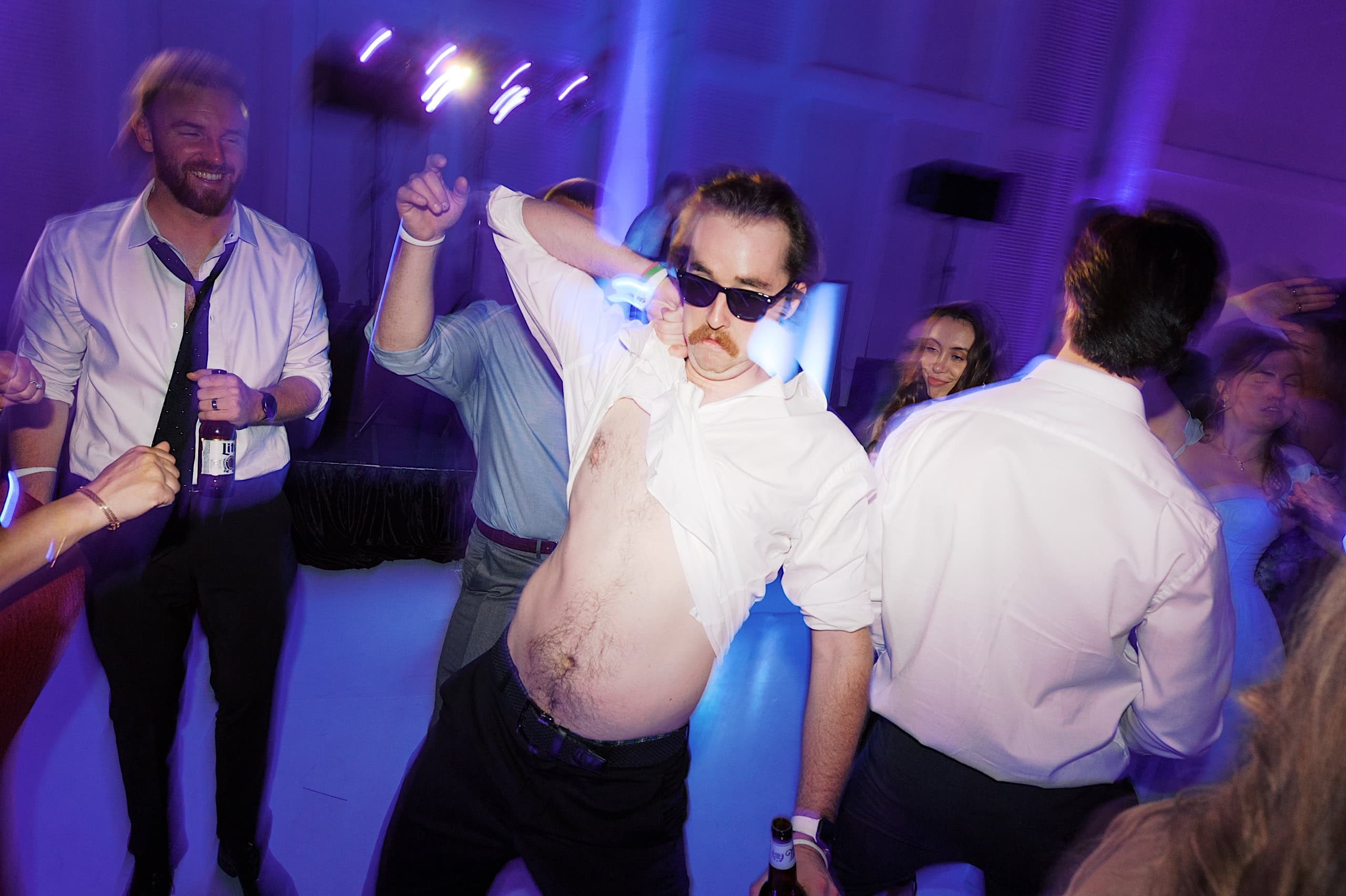 A man in sunglasses lifts his shirt and dances energetically at a party, surrounded by others in dress shirts under purple lighting, celebrating after a joyful The Ivy at Ellis Preserve spring wedding.