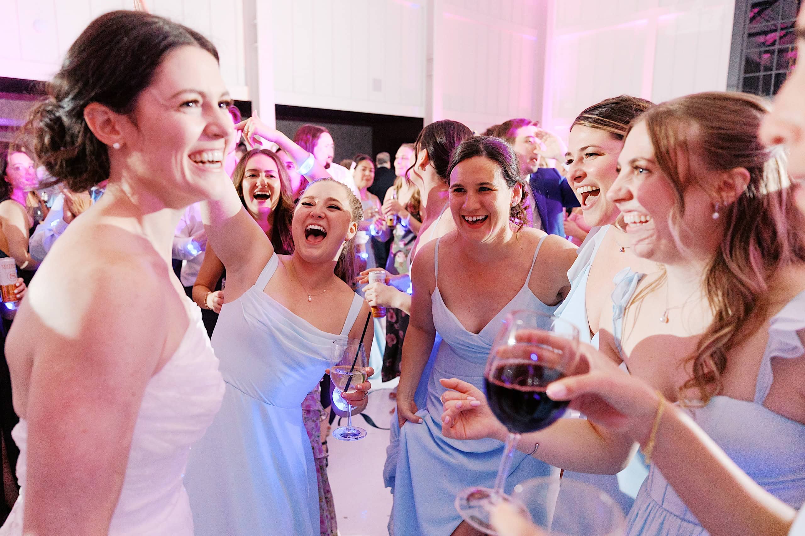 A bride in a white dress smiles and celebrates with bridesmaids in light blue dresses at an indoor The Ivy at Ellis Preserve spring wedding reception.