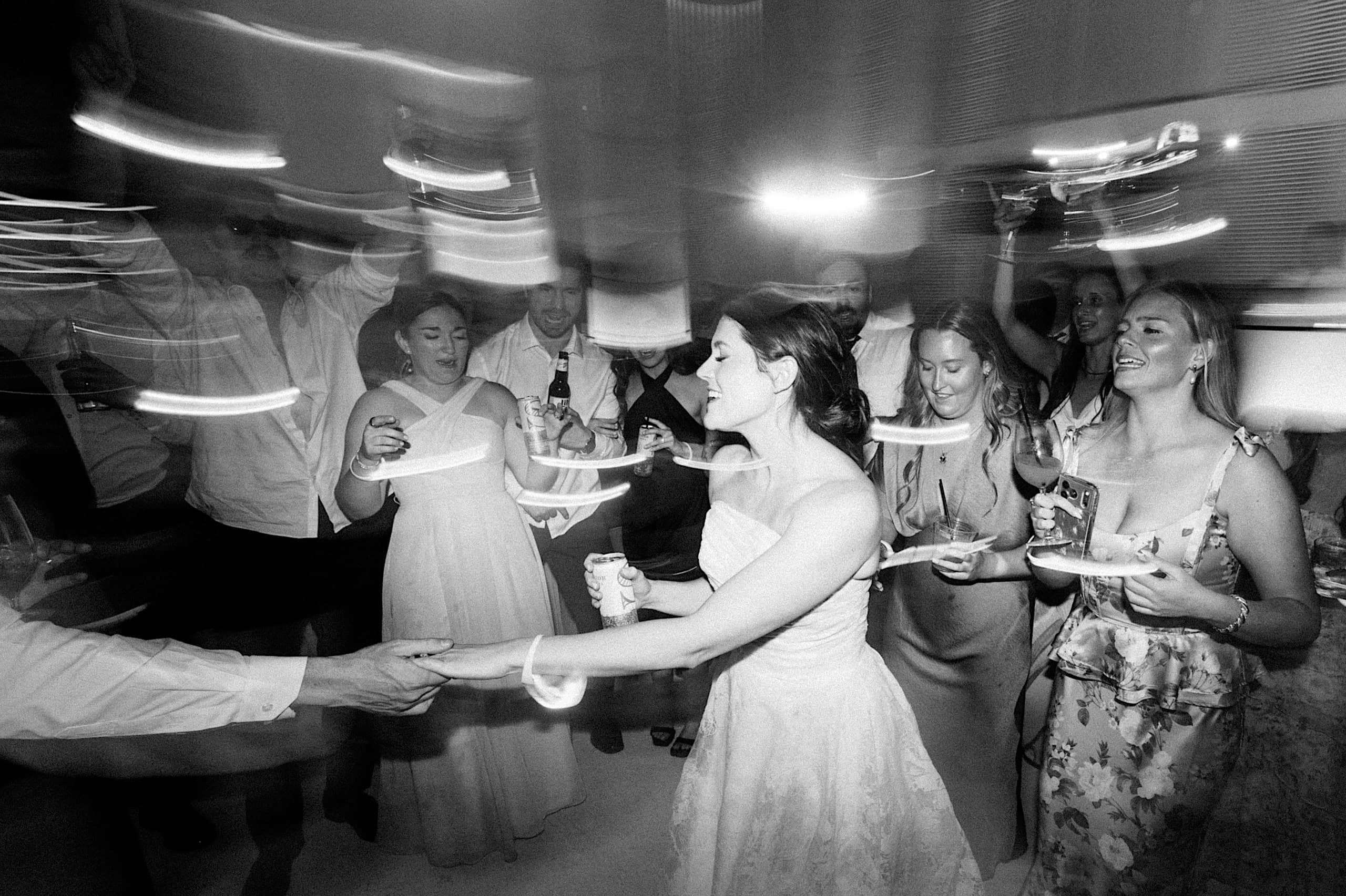 A bride in a wedding dress dances with guests around her, holding drinks and plates, under dynamic lighting at a lively indoor The Ivy at Ellis Preserve spring wedding celebration.