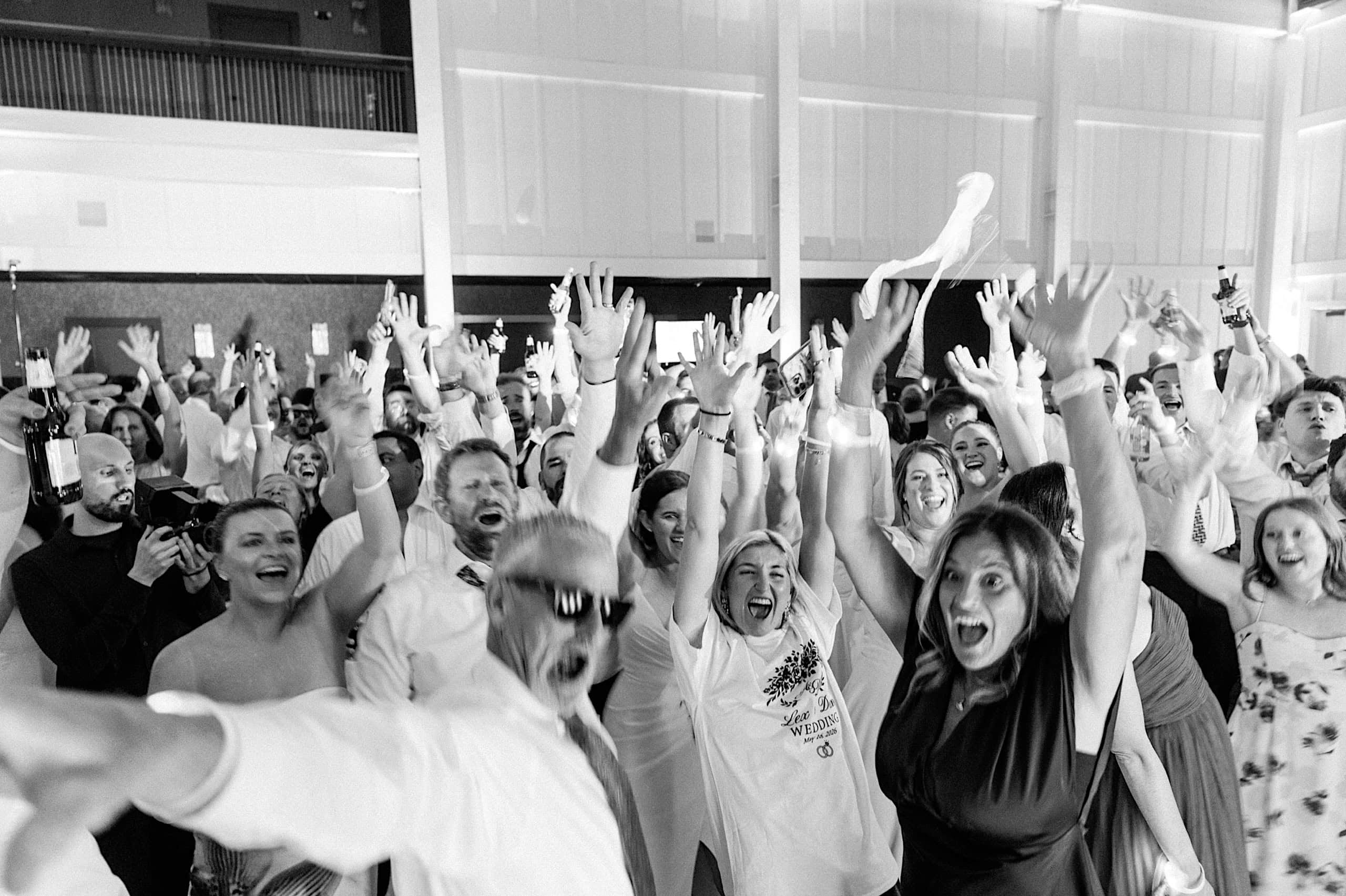 A large group of people indoors raise their hands and cheer energetically, with many smiling at the camera during a lively, festive celebration at The Ivy at Ellis Preserve spring wedding.