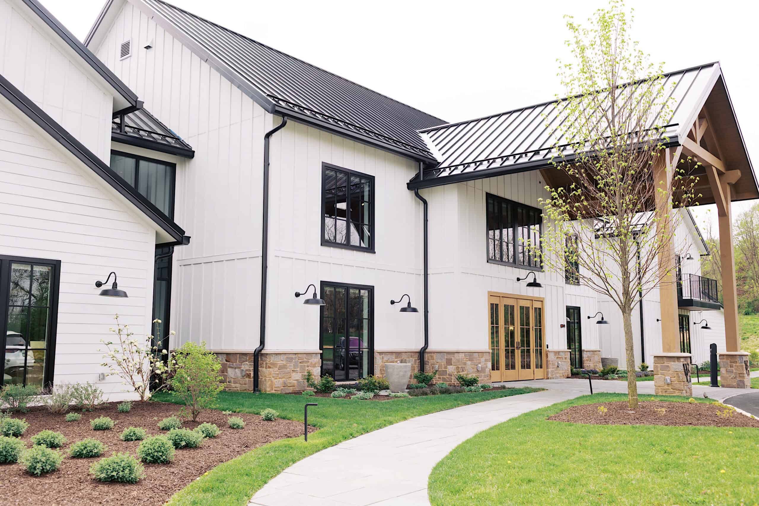 Modern white barn-style building with black trim, stone accents, large windows, and double wooden doors—perfect for a Westwynd Gardens Spring Wedding—surrounded by landscaped greenery and a curved sidewalk.