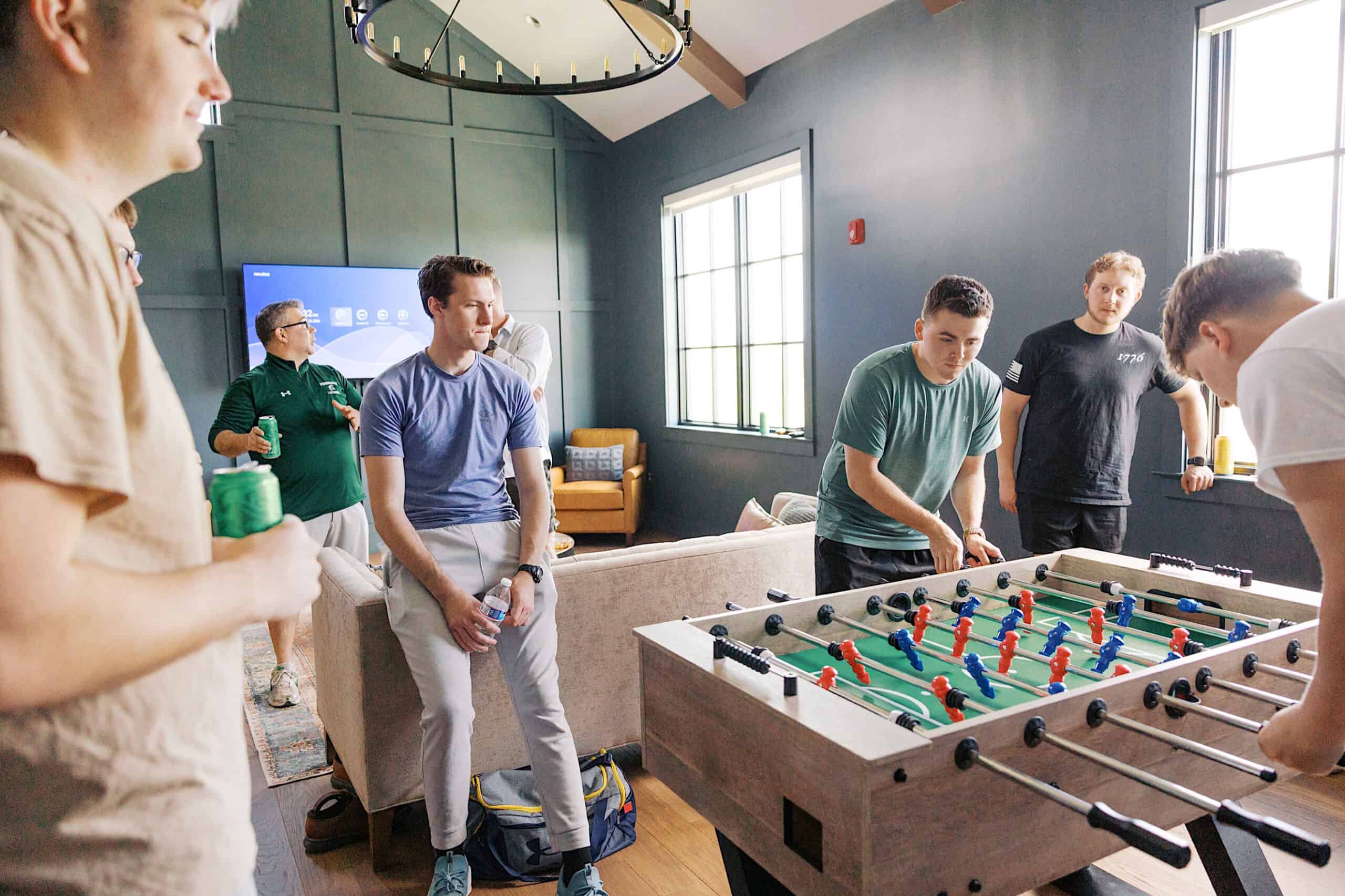 A group of people in a casual room watch two individuals play foosball near a couch and large windows, echoing the lively camaraderie often found at a Westwynd Gardens Spring Wedding.