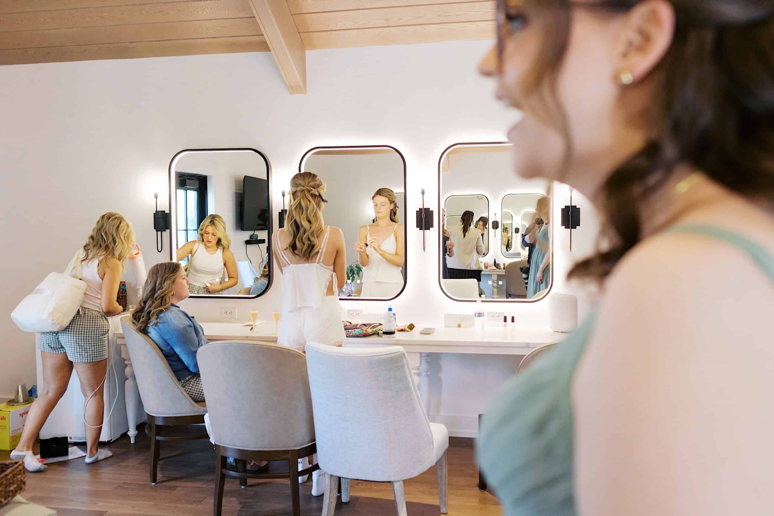 Four women gather in a brightly lit dressing room at a Westwynd Gardens Spring Wedding; three are by the vanity with mirrors while one stands in the foreground, partially out of focus.
