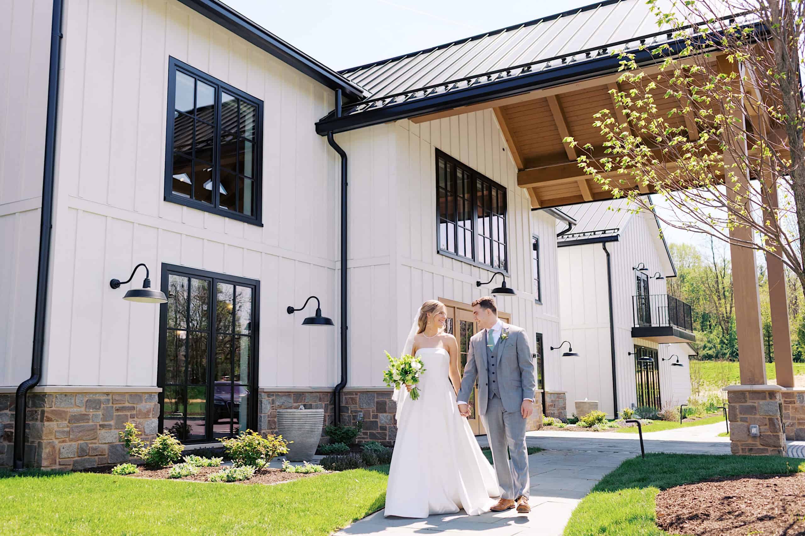 A bride and groom in wedding attire walk hand in hand outside a modern barn-style building at Westwynd Gardens, soaking up the sunshine on their beautiful spring wedding day.