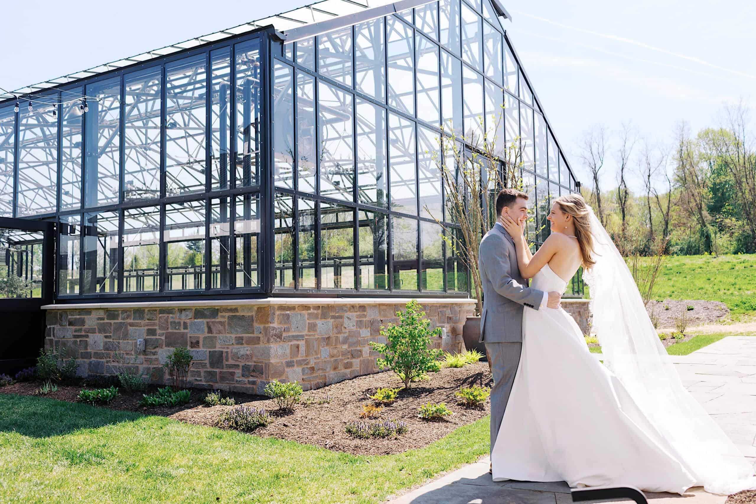 A bride and groom stand outside the modern glass greenhouse at Westwynd Gardens on a sunny day, holding each other and smiling during their beautiful spring wedding.