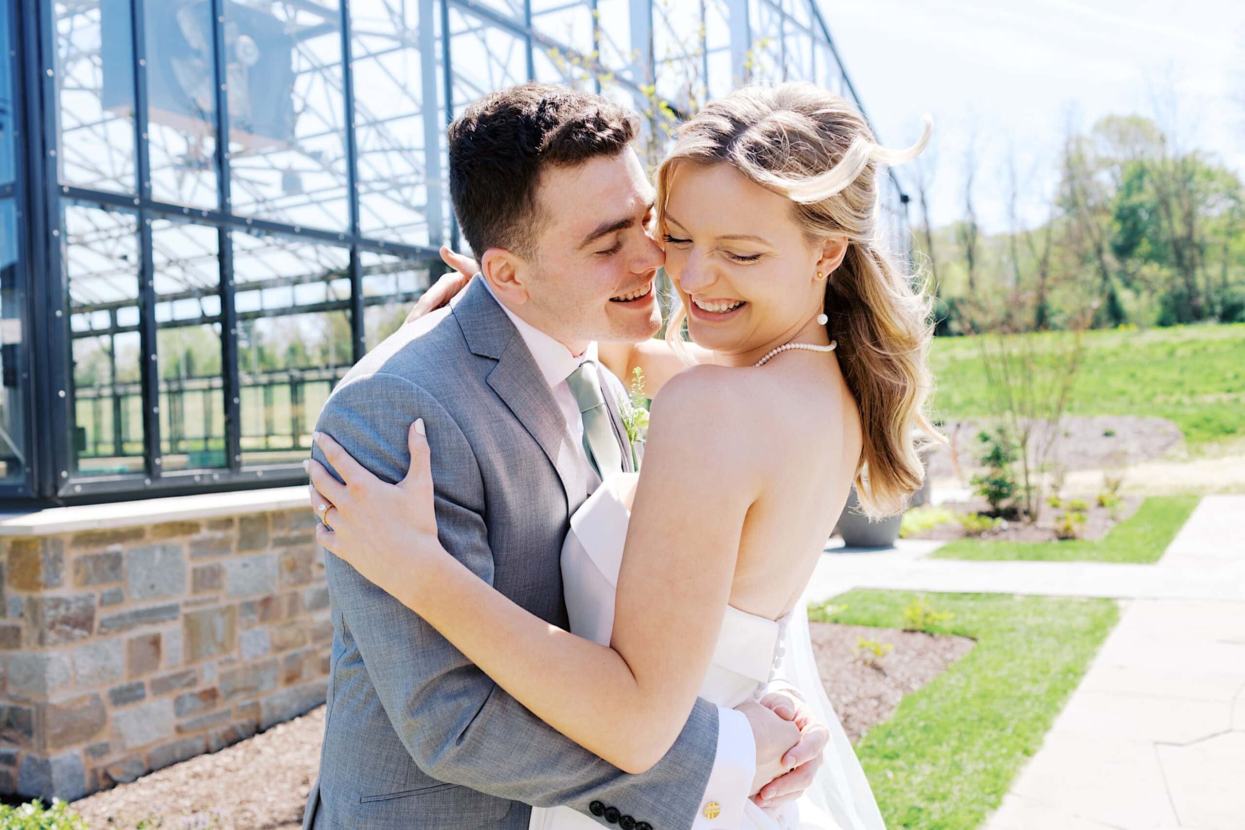 A bride and groom embrace and smile outdoors on a sunny day at their Westwynd Gardens Spring Wedding, near a glass building and landscaped garden.