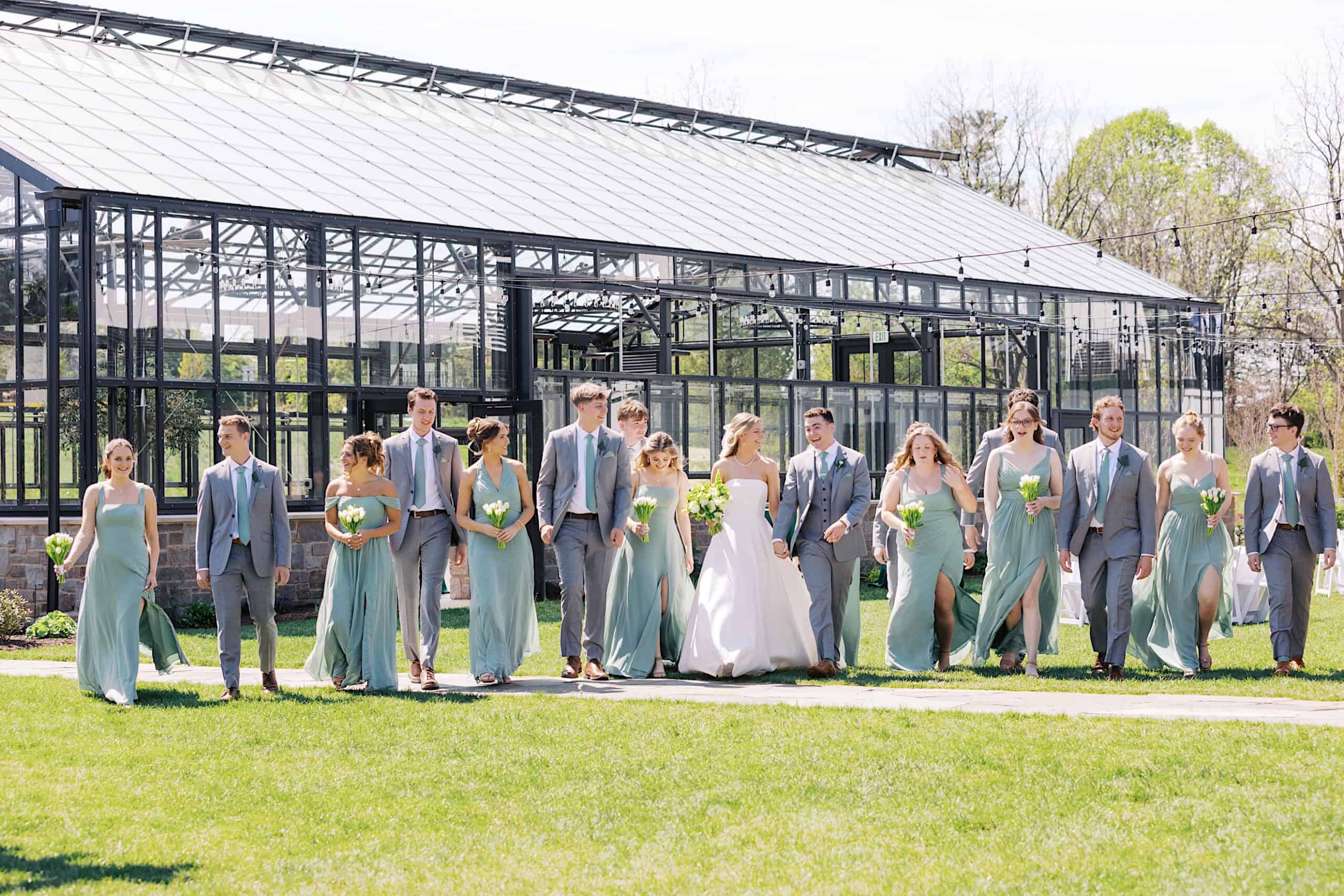 A wedding party in pastel attire walks together on grass outside the large glass greenhouse at Westwynd Gardens, celebrating a joyful Spring Wedding on a sunny day.
