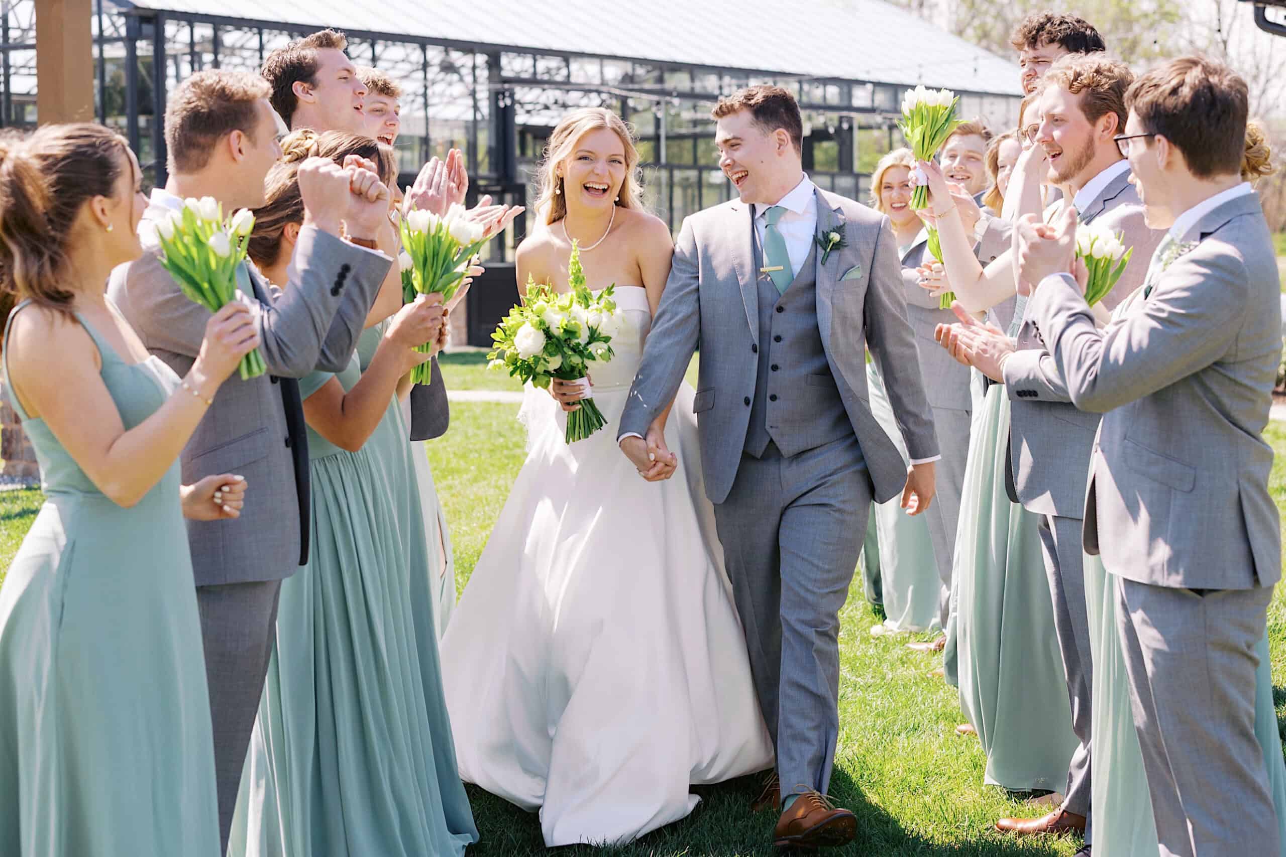 A bride and groom walk outdoors at their Westwynd Gardens Spring Wedding, smiling and holding hands, as their wedding party in matching outfits cheers and holds bouquets.