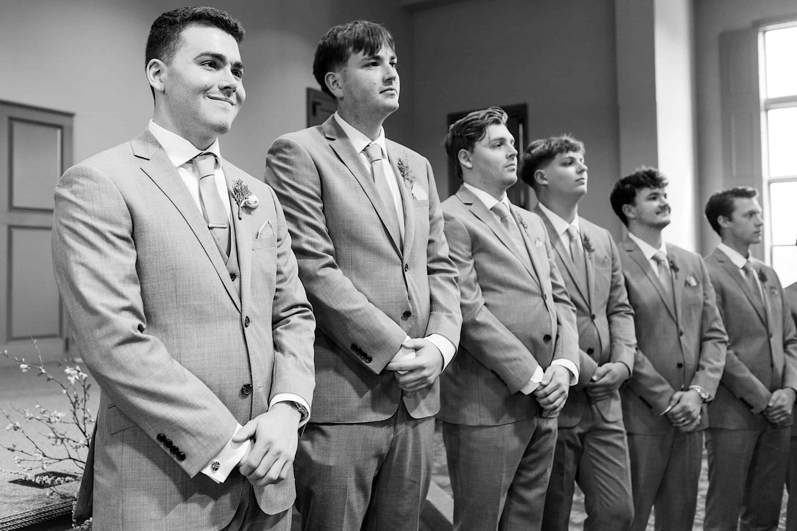 Six men in light-colored suits stand in a row indoors at a Westwynd Gardens Spring Wedding, facing forward with hands clasped, appearing to participate in a formal event.