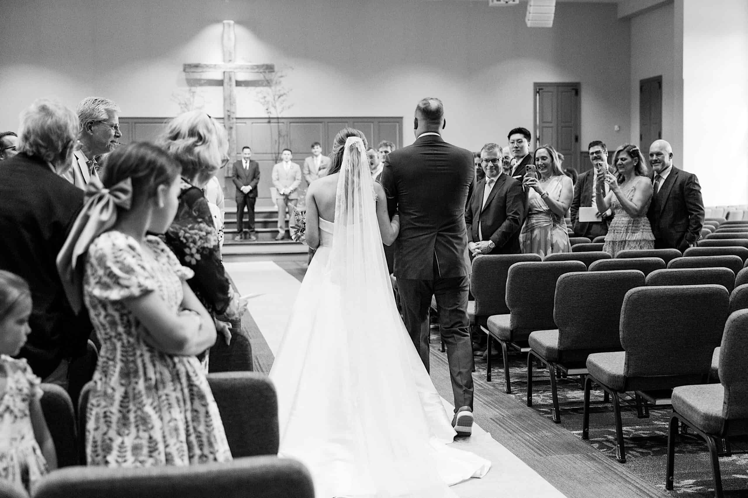 A bride and groom walk down the aisle together in a church at their Westwynd Gardens Spring Wedding as guests stand and watch; a cross is visible at the front.