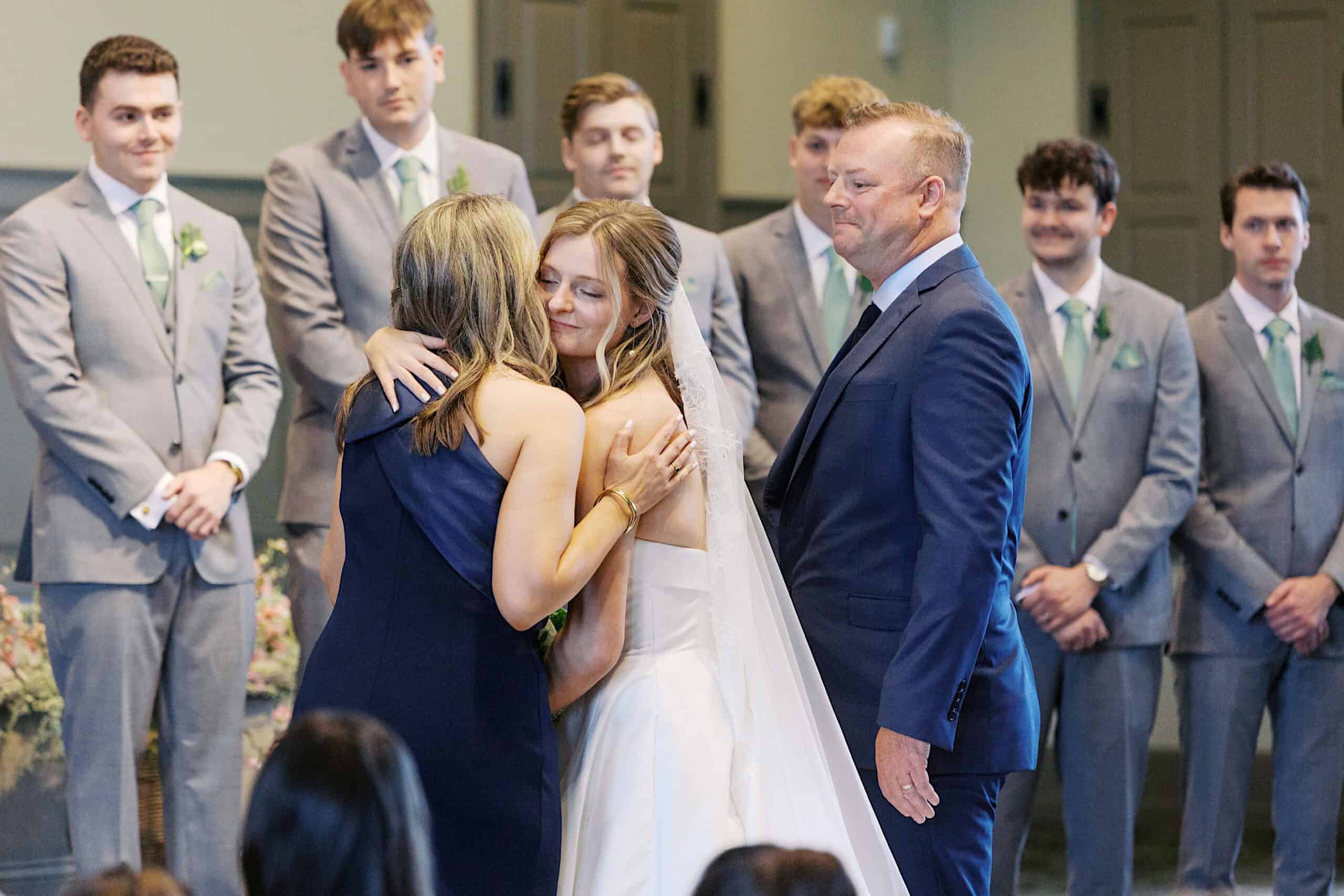 At a Westwynd Gardens Spring Wedding, a bride in a white dress hugs a woman while a man in a suit stands beside them; groomsmen in gray suits look on in the background during the ceremony.