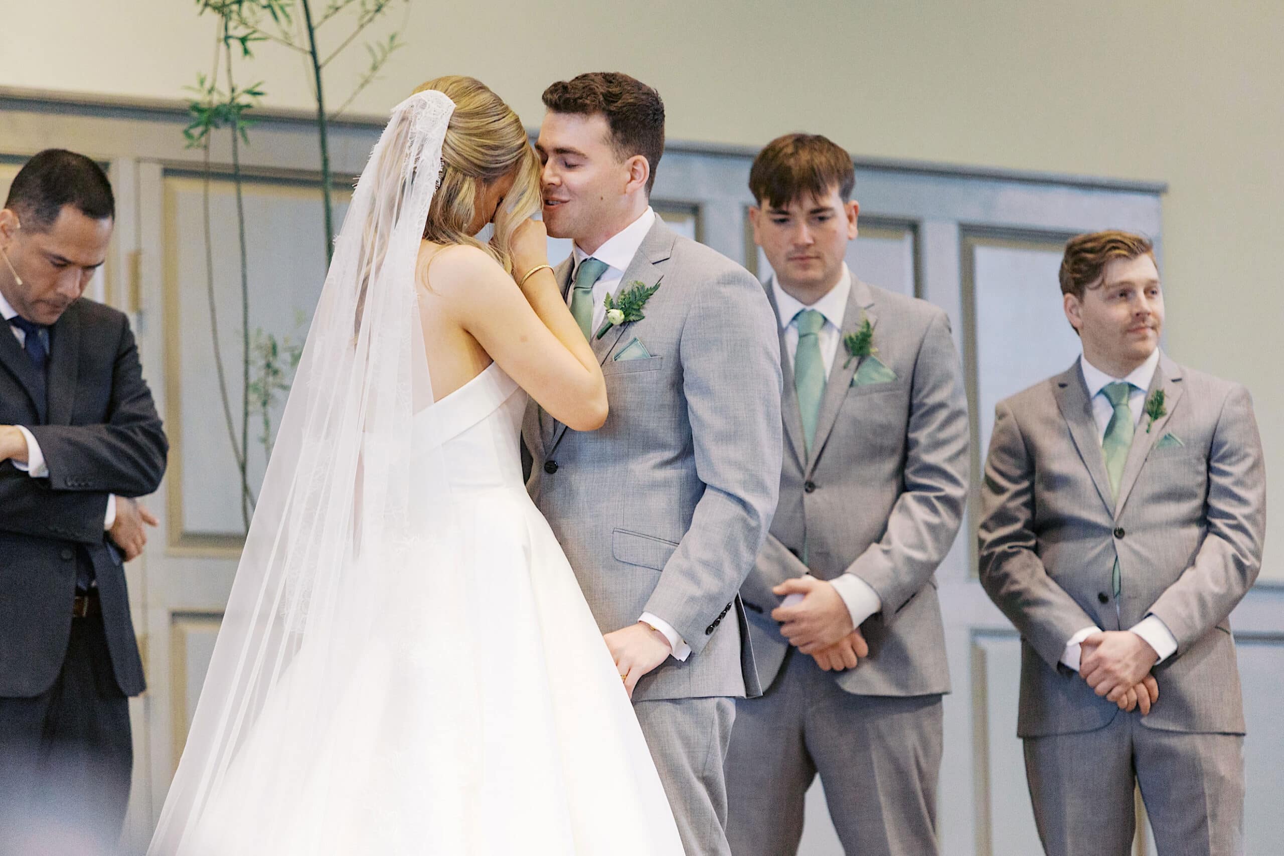 A bride and groom share a moment during their Westwynd Gardens Spring Wedding, standing together in front of groomsmen in gray suits.