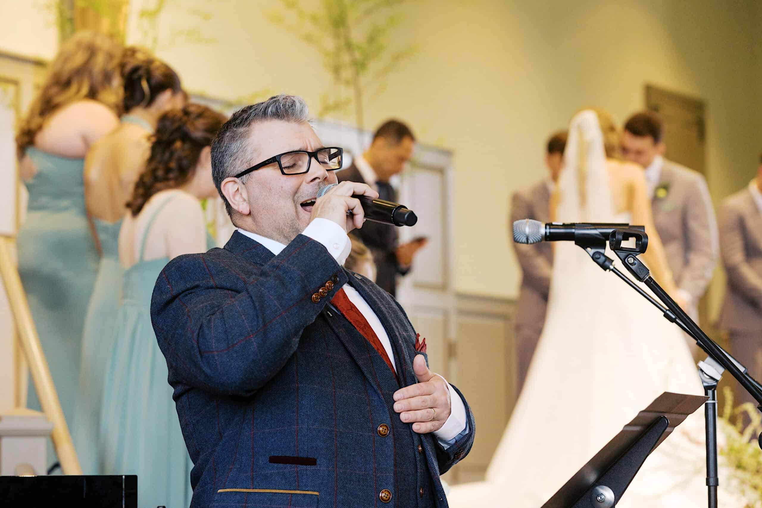 At a Westwynd Gardens Spring Wedding, a man in a suit sings into a microphone during the ceremony, with the bride, groom, and wedding party elegantly gathered in the background.