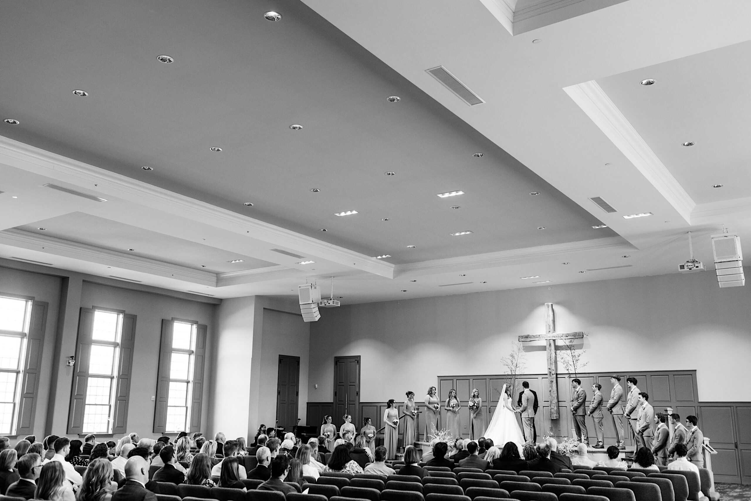 A Westwynd Gardens Spring Wedding ceremony unfolds in a church, with the bride, groom, wedding party, and guests seated; a large cross is displayed on the wall behind the couple.