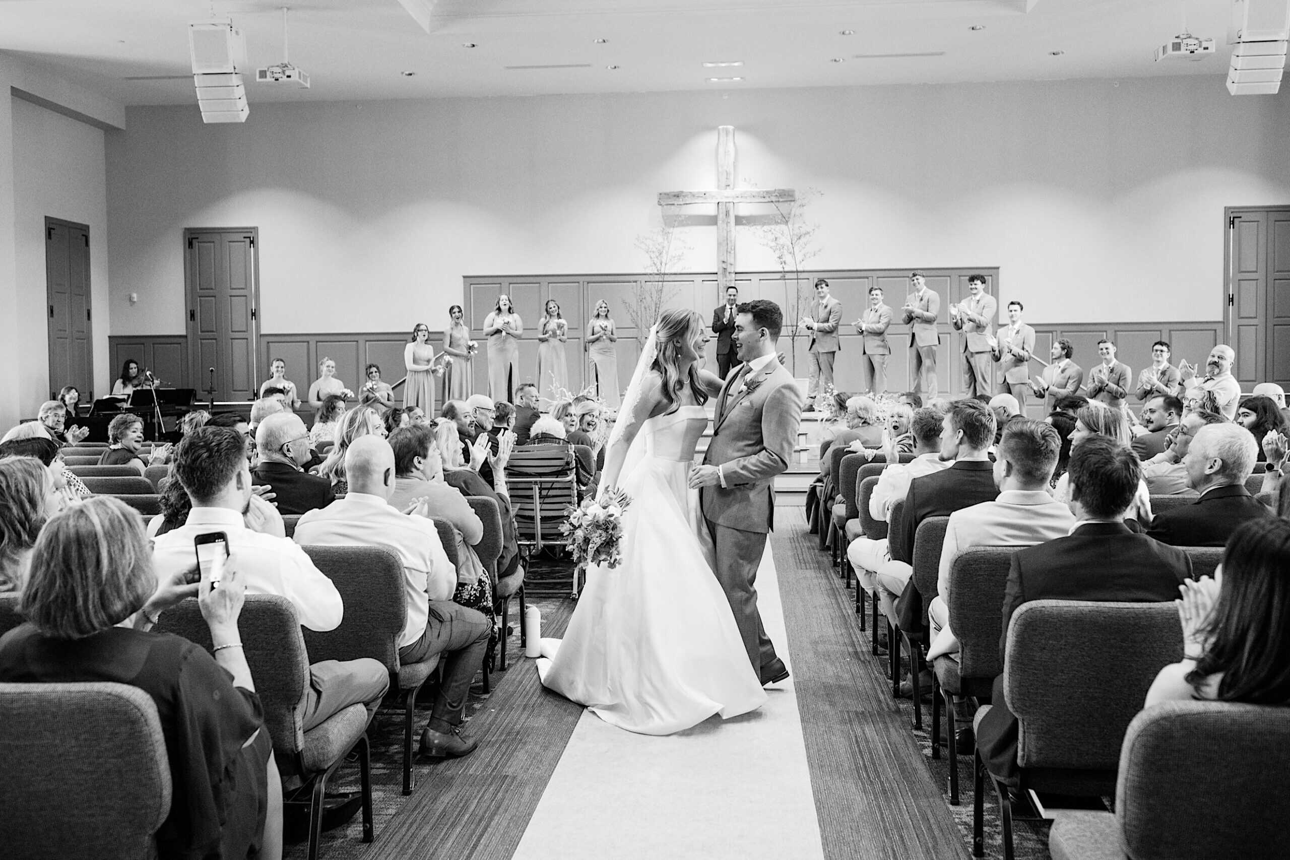 A bride and groom share a kiss at the end of the aisle during their Westwynd Gardens Spring Wedding, surrounded by seated guests and their wedding party onstage.