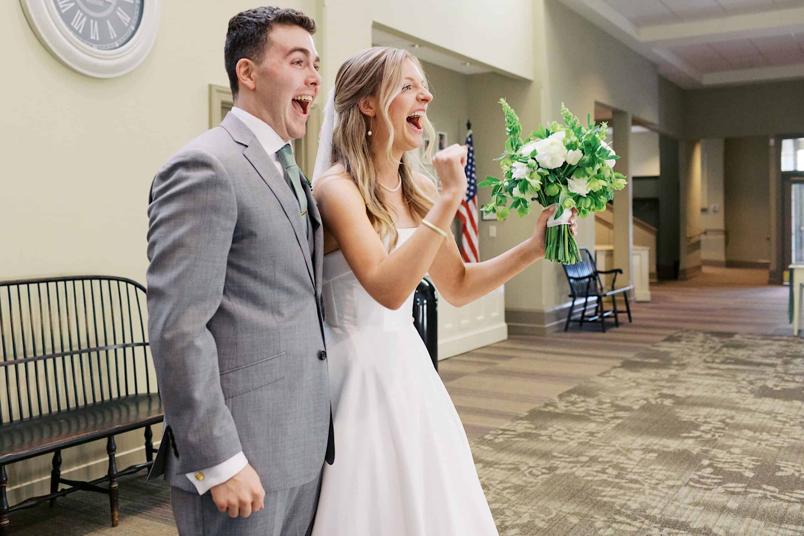 A bride and groom stand indoors at their Westwynd Gardens Spring Wedding, smiling and cheering. The bride holds a bouquet of white flowers, and both appear excited. An American flag and large clock are visible in the background.