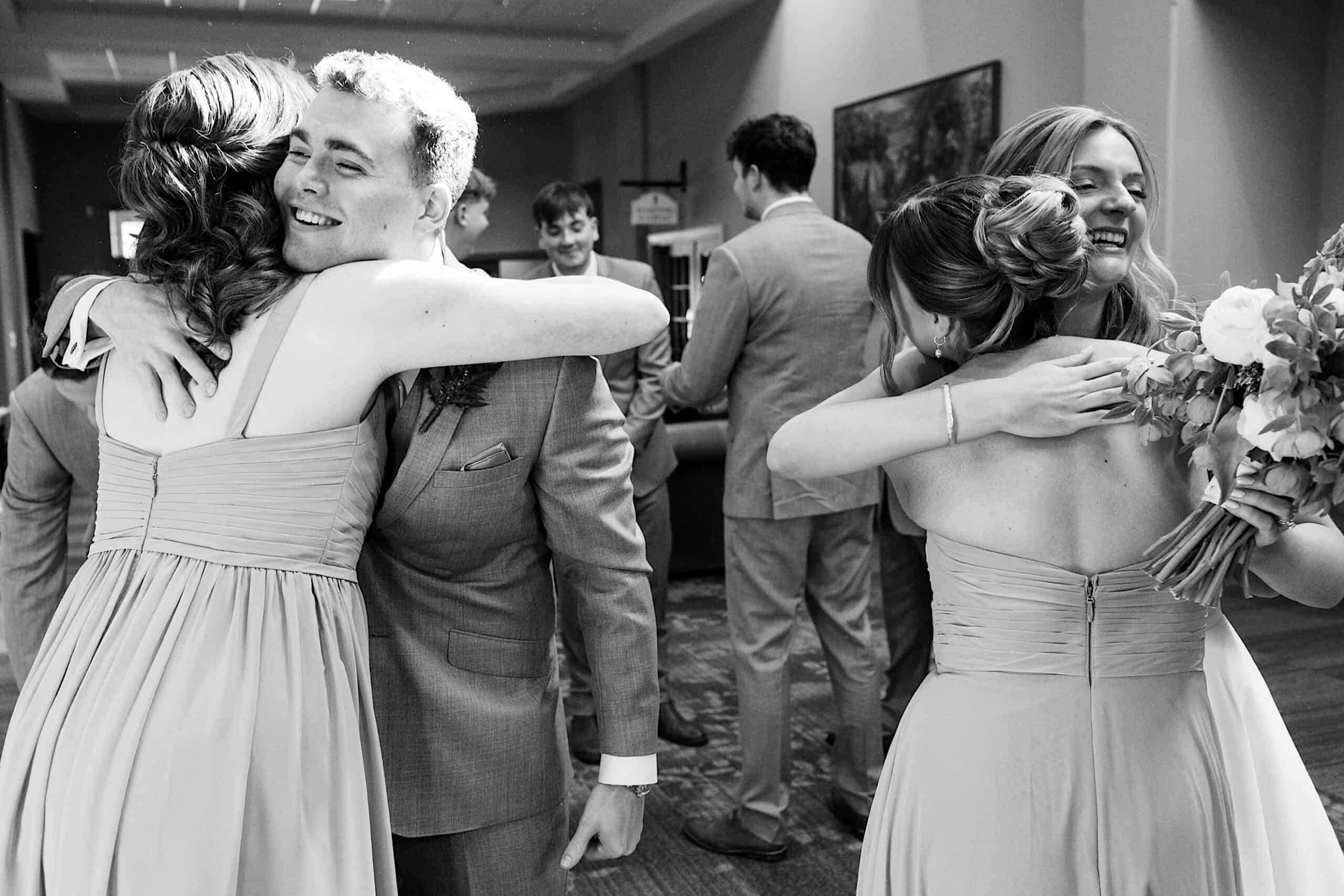 A group of people in formal attire exchange hugs and smiles indoors at a Westwynd Gardens Spring Wedding, with two women in dresses and two men in suits in the foreground.