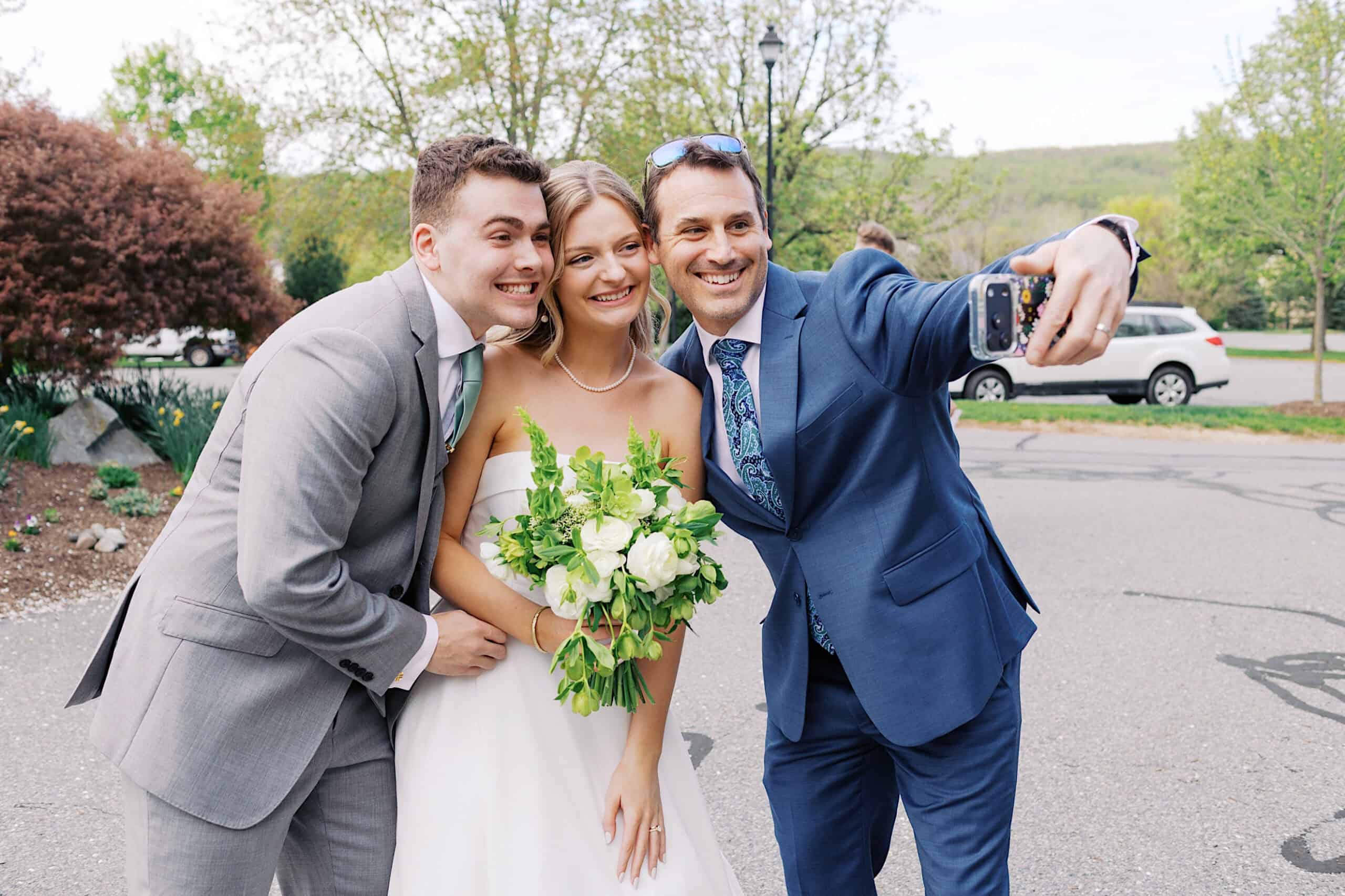 Three people dressed formally, including a bride holding a bouquet, pose together outdoors at a Westwynd Gardens Spring Wedding as one person takes a selfie with a smartphone.