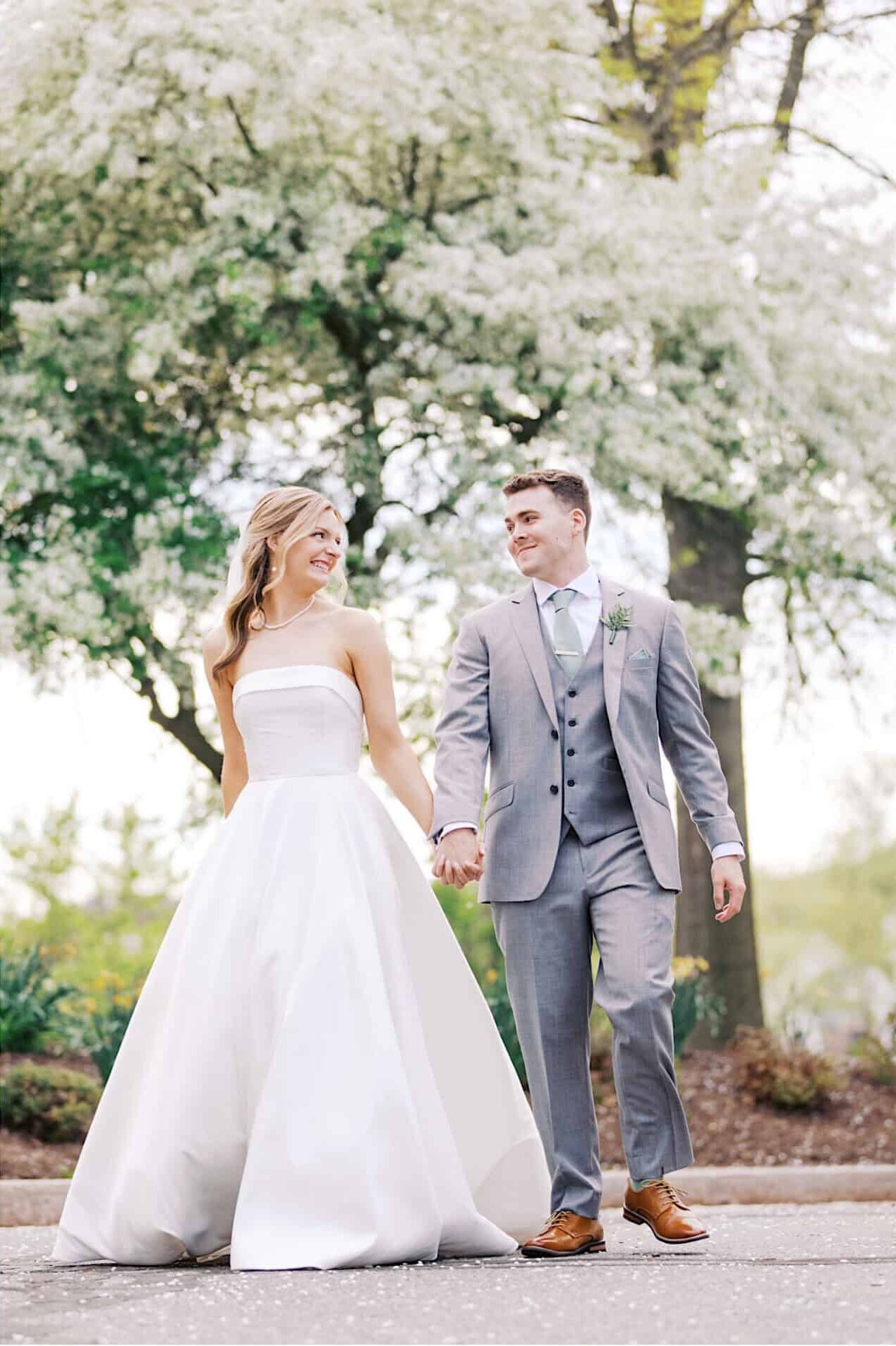 A bride in a white dress and a groom in a gray suit share a kiss and walk hand in hand outdoors, surrounded by blooming trees during their Westwynd Gardens Spring Wedding.