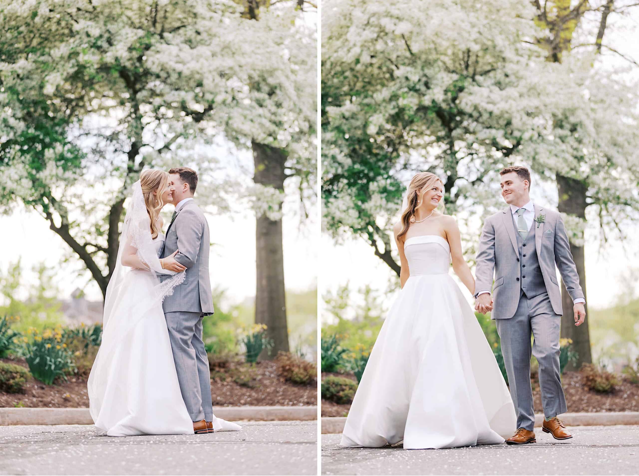 A bride in a white dress and a groom in a gray suit share a kiss and walk hand in hand outdoors, surrounded by blooming trees during their Westwynd Gardens Spring Wedding.