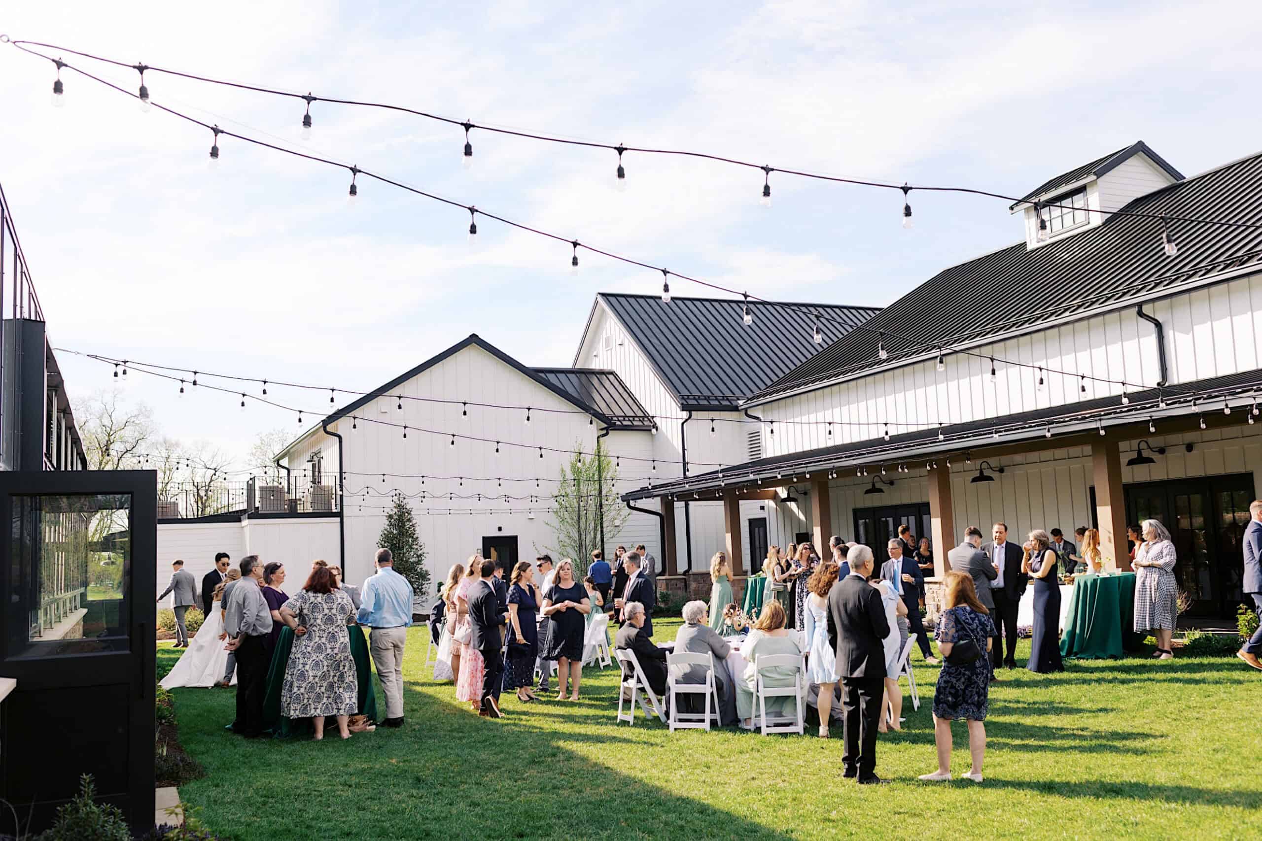 A group of people gather outdoors on a lawn for a Westwynd Gardens Spring Wedding, with string lights overhead and barn-style buildings in the background under a clear sky.