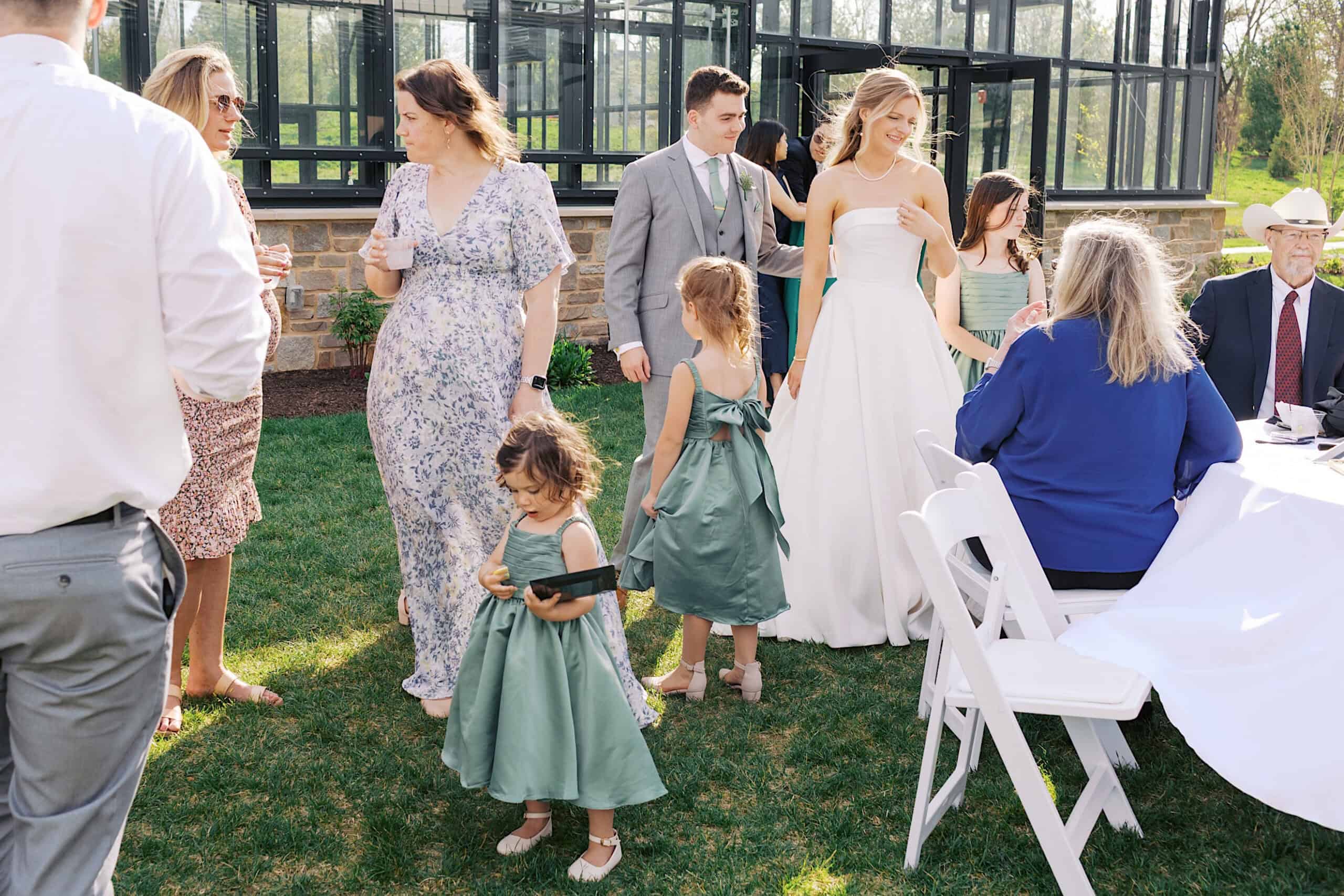 A bride and groom stand outside with guests, including children in green dresses, at a Westwynd Gardens Spring Wedding near a glass greenhouse.