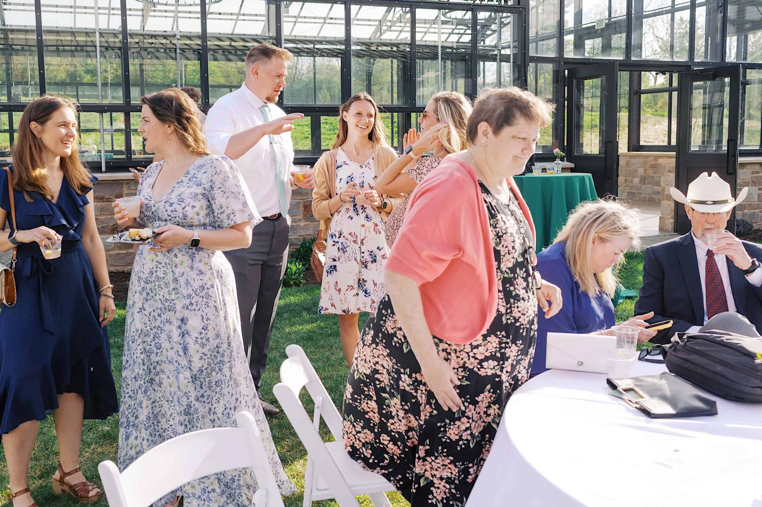 A group of people, some standing and some seated, gather outdoors near white tables and chairs at a Westwynd Gardens Spring Wedding, with a glass greenhouse in the background on a sunny day.