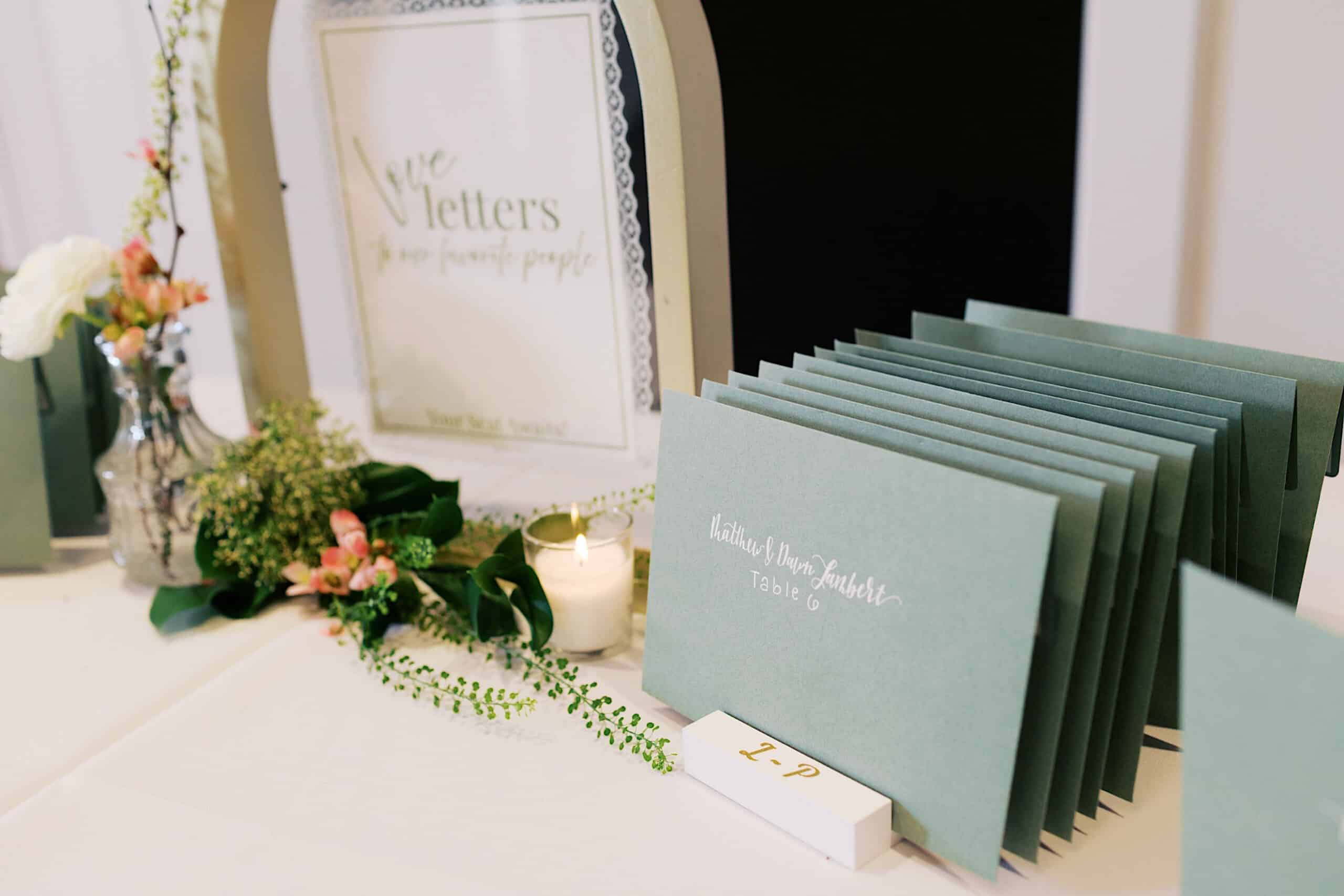 A display of green envelopes labeled with names and table numbers, a framed sign, flowers, a candle, and a small white box creates an inviting welcome at this Westwynd Gardens Spring Wedding.