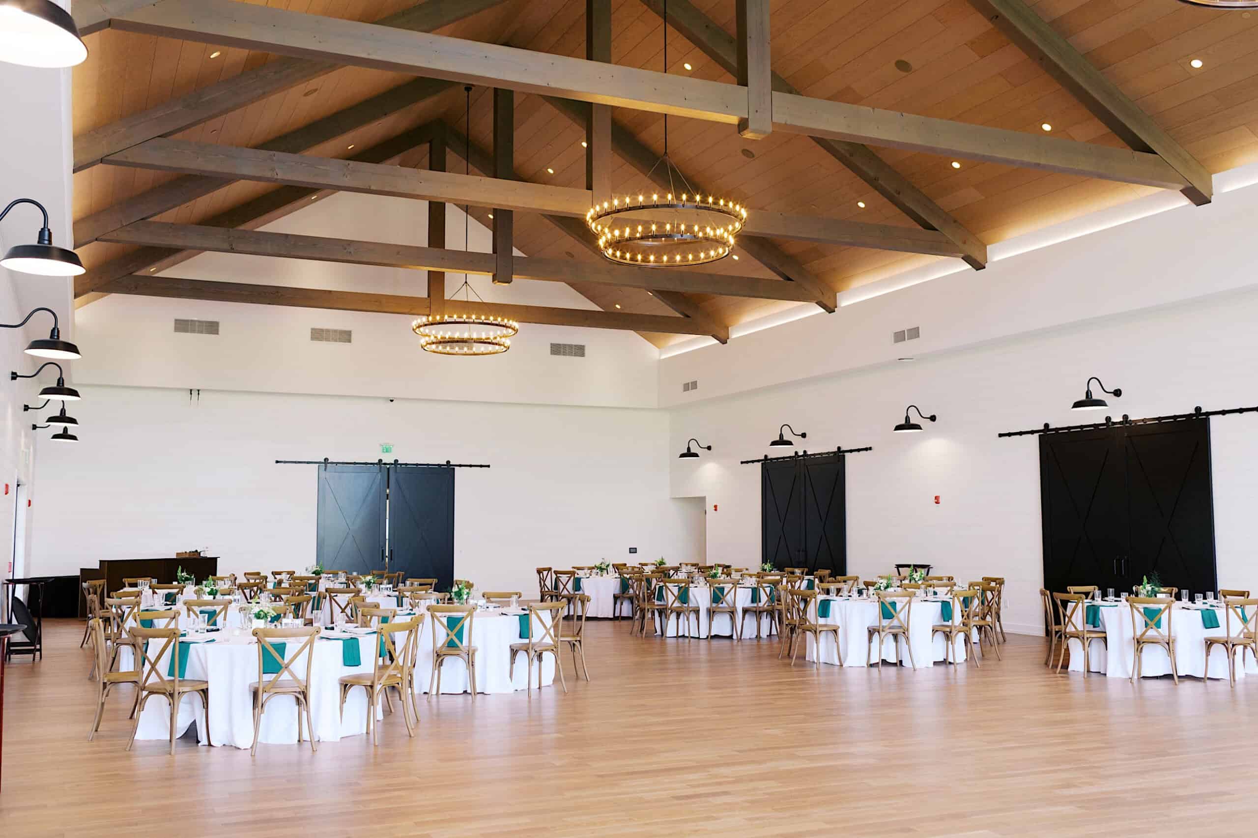 A spacious event hall at Westwynd Gardens, perfect for a Spring Wedding, features high wooden ceilings, circular chandeliers, and round tables covered in white tablecloths arranged for a beautiful gathering.