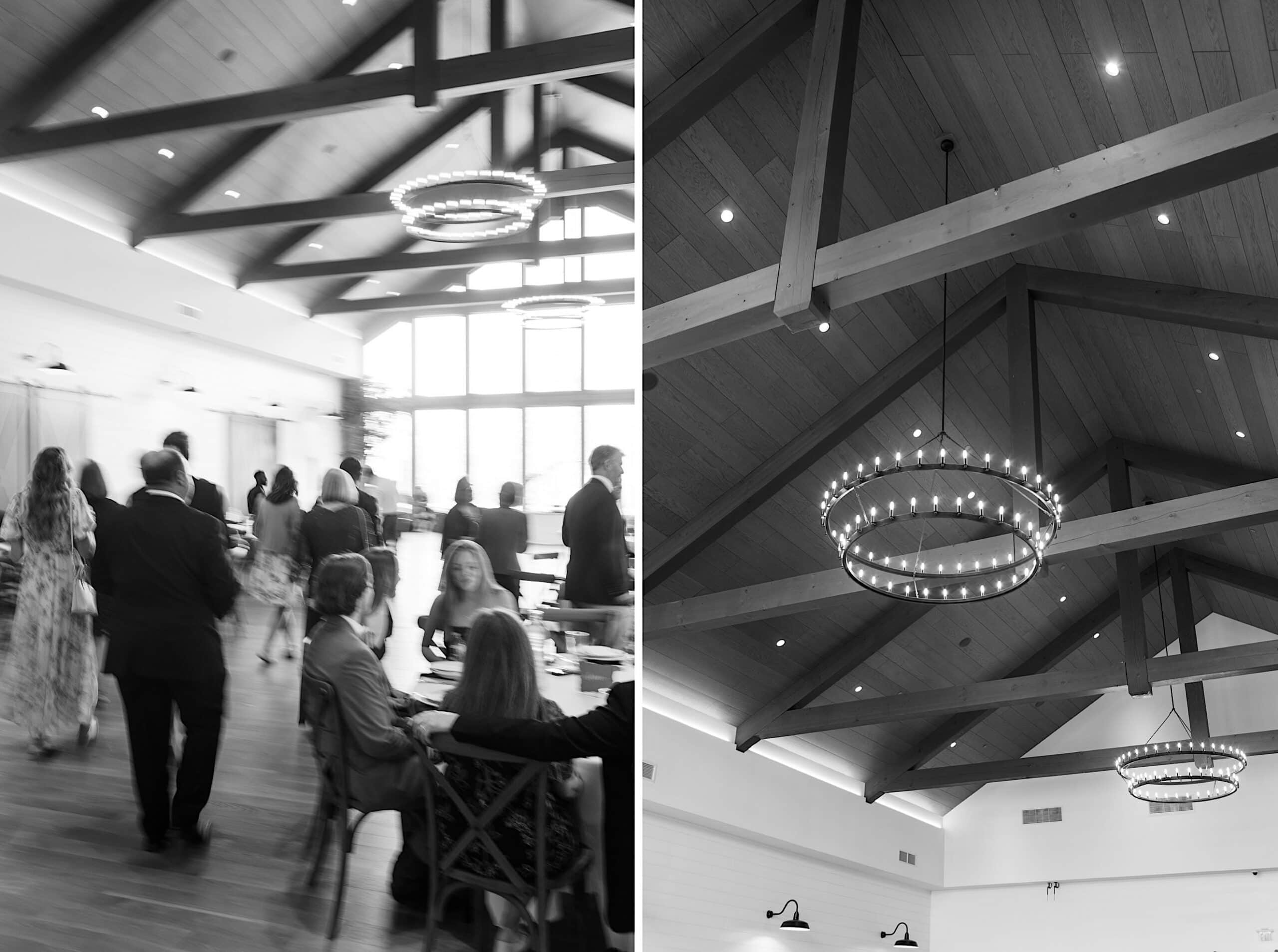 Black and white photo of an indoor event space at a Westwynd Gardens Spring Wedding, with exposed wooden beams, round chandeliers, and people gathered—some seated, others standing or walking.