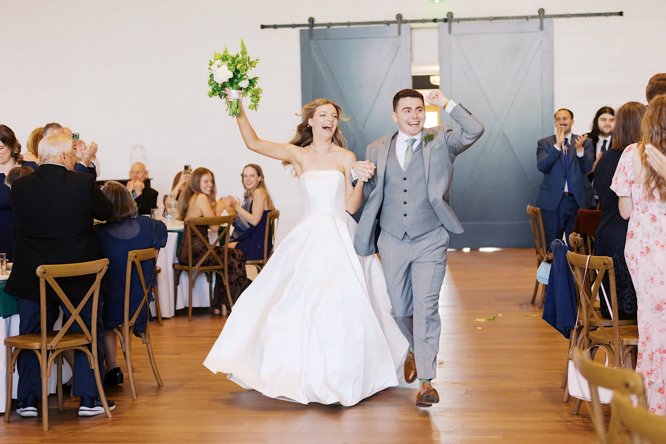 A bride and groom, holding hands and smiling, walk joyfully into their Westwynd Gardens Spring Wedding reception as guests applaud.