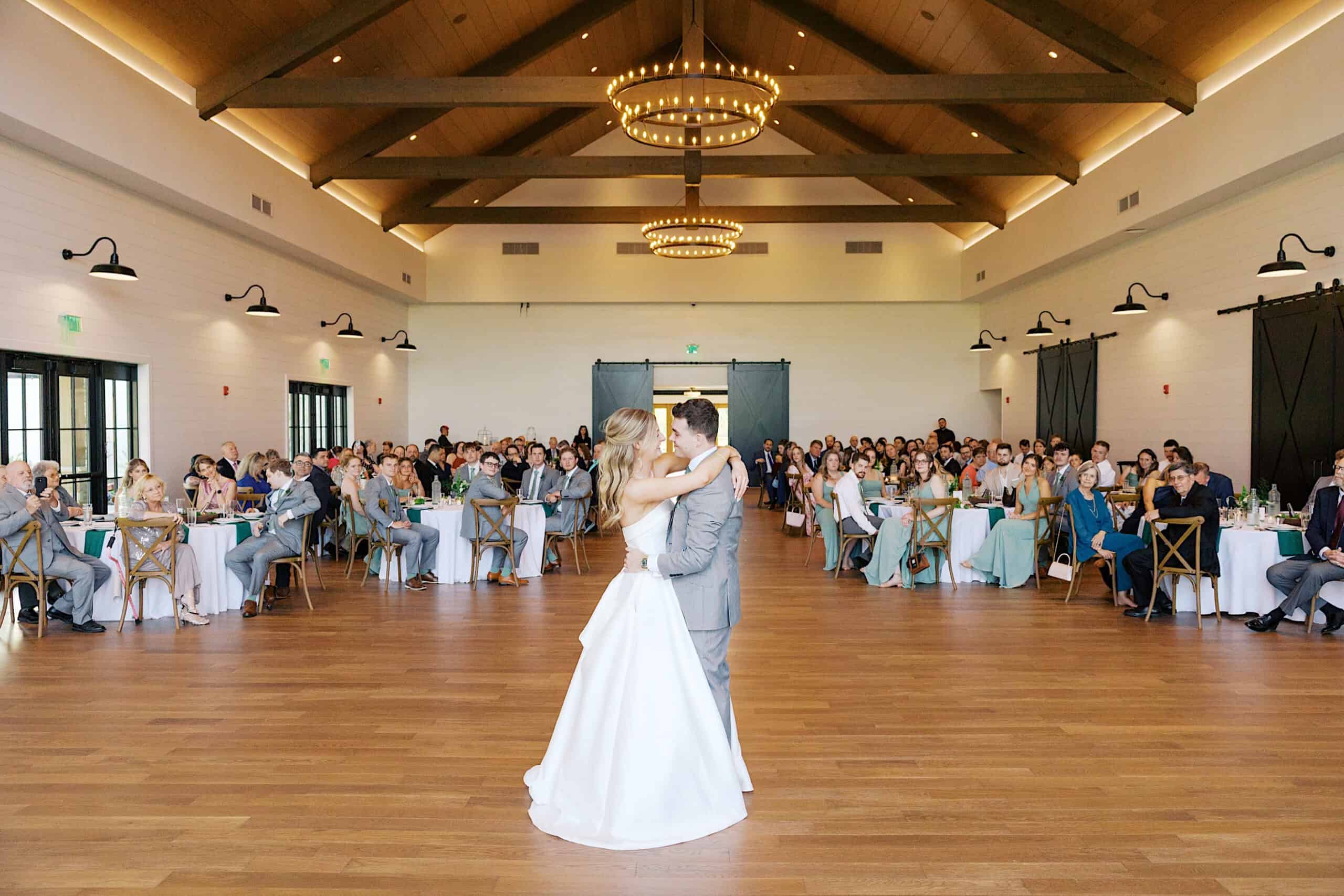A bride and groom share their first dance in a spacious, well-lit reception hall at a Westwynd Gardens Spring Wedding, with guests seated at round tables watching.