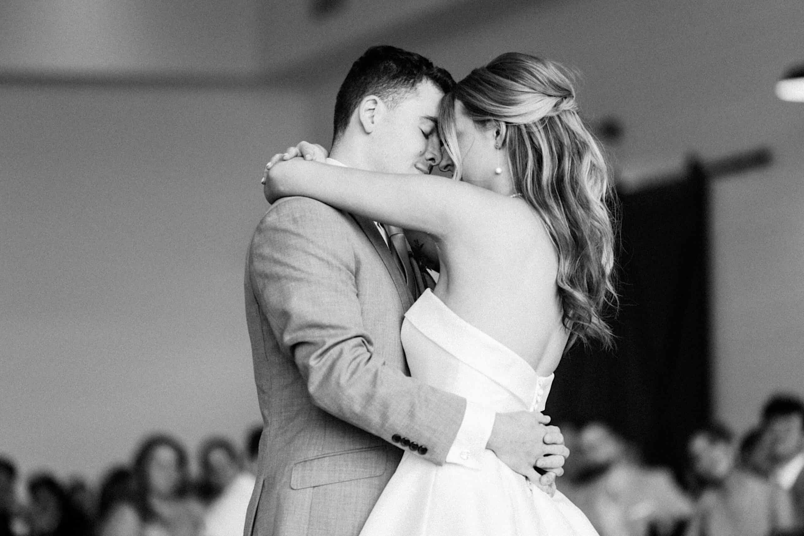 A couple dressed in formal attire shares a close embrace while dancing at an indoor event during a Westwynd Gardens Spring Wedding, with a blurred crowd in the background.