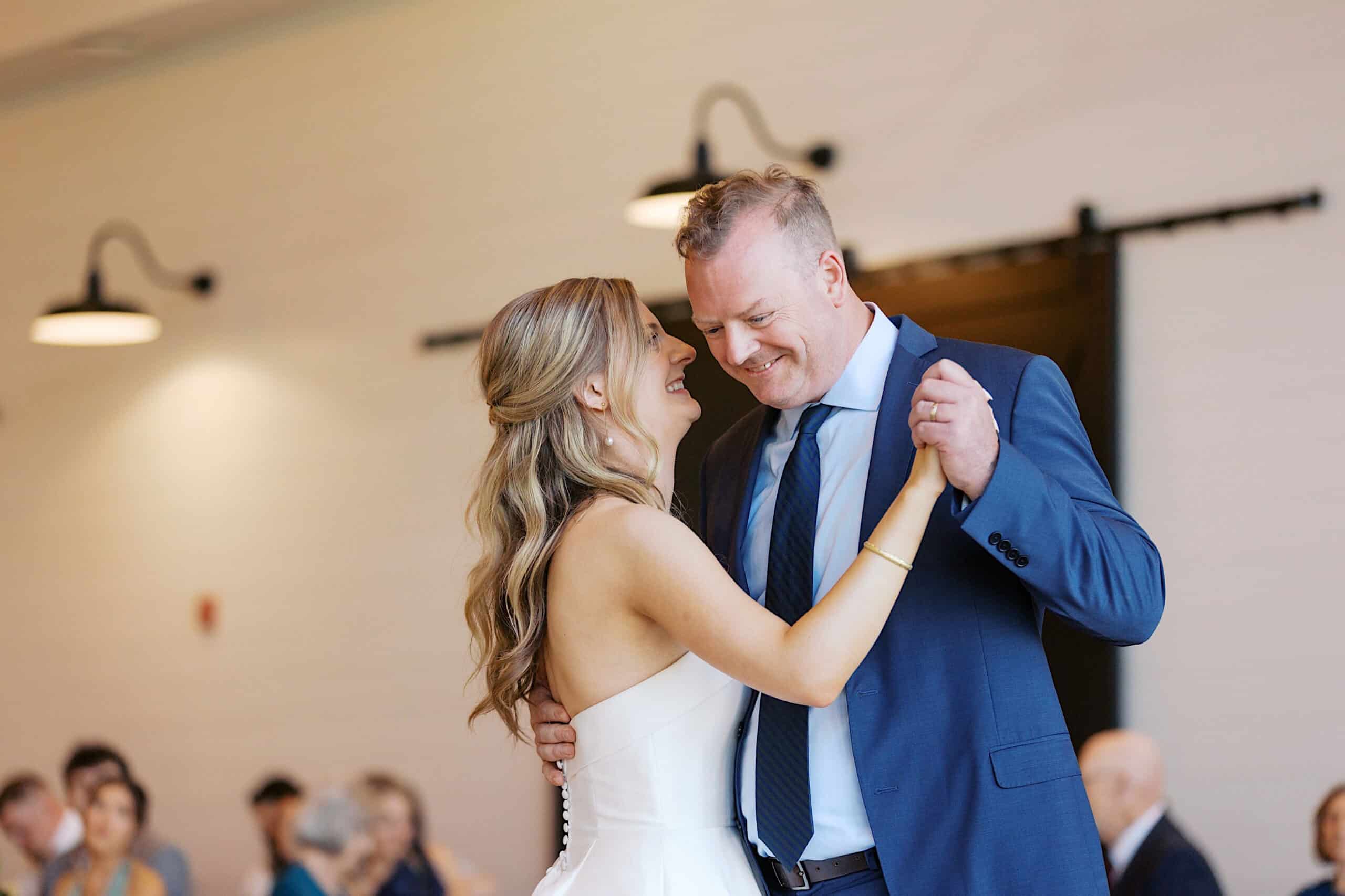 A bride in a white dress and a man in a blue suit smile while dancing together at their Westwynd Gardens Spring Wedding, with guests seated in the background.