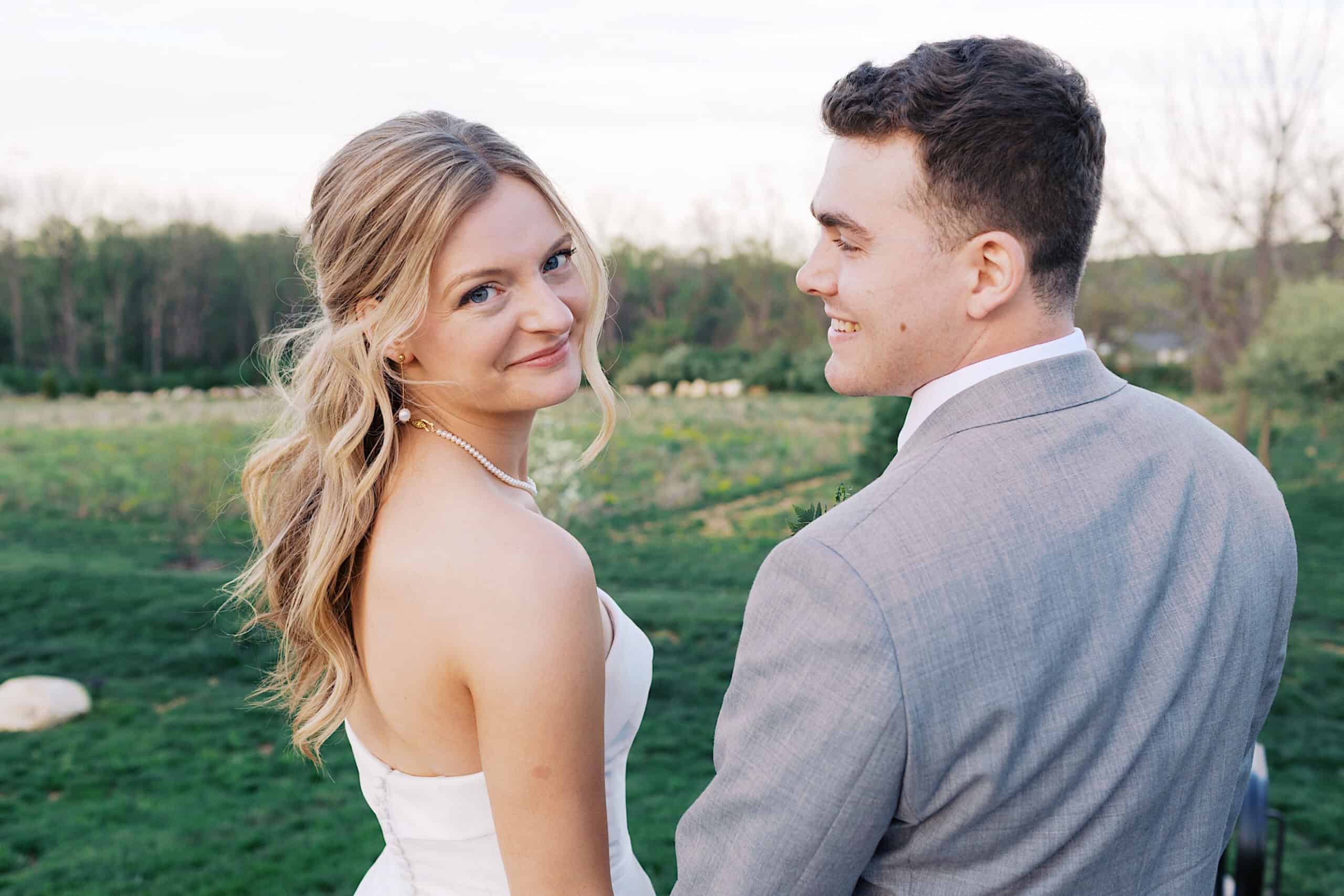 A bride and groom stand outdoors on grass at their Westwynd Gardens Spring Wedding, holding hands and smiling back at the camera, with trees and lush greenery in the background.