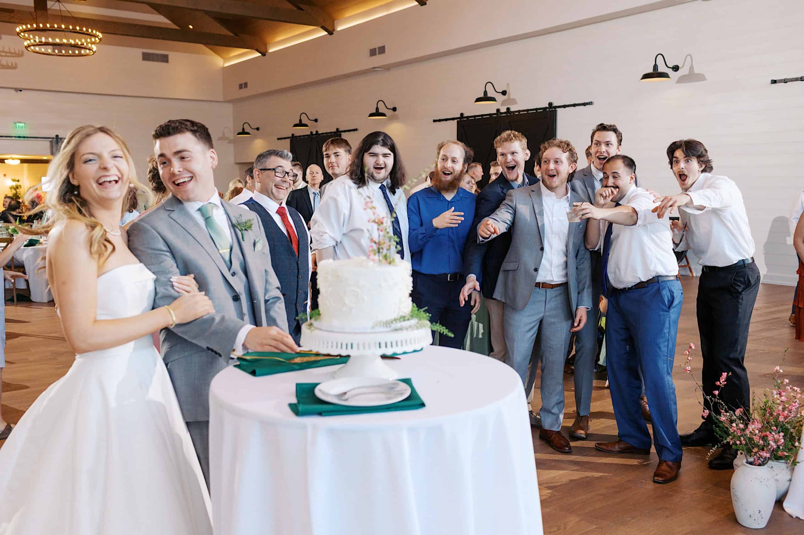 A bride and groom stand by a wedding cake, smiling, while guests in formal attire react excitedly in the background at their Westwynd Gardens Spring Wedding.