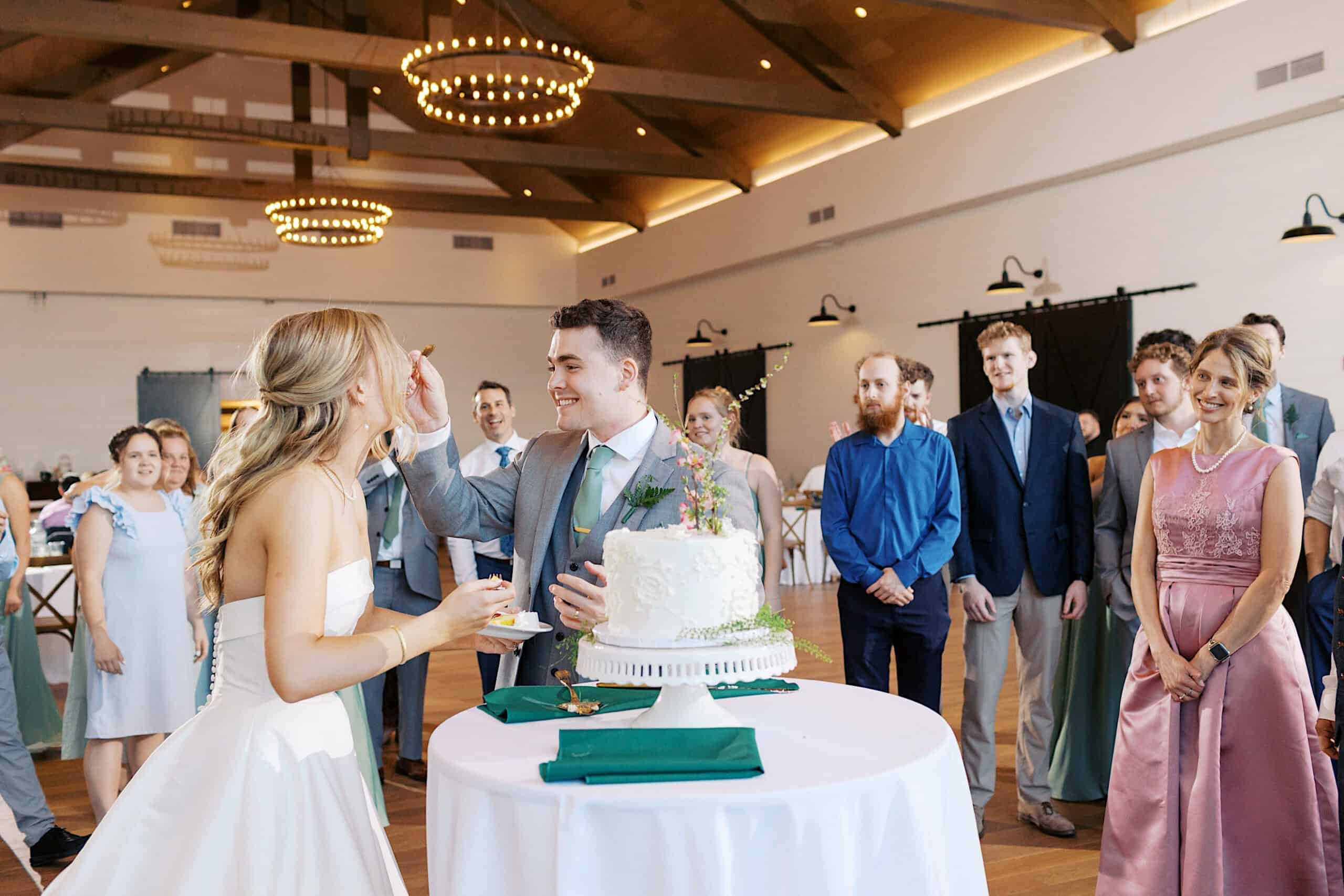 At a Westwynd Gardens Spring Wedding, a bride feeds cake to the groom at the reception while guests watch and smile inside the beautifully decorated hall.