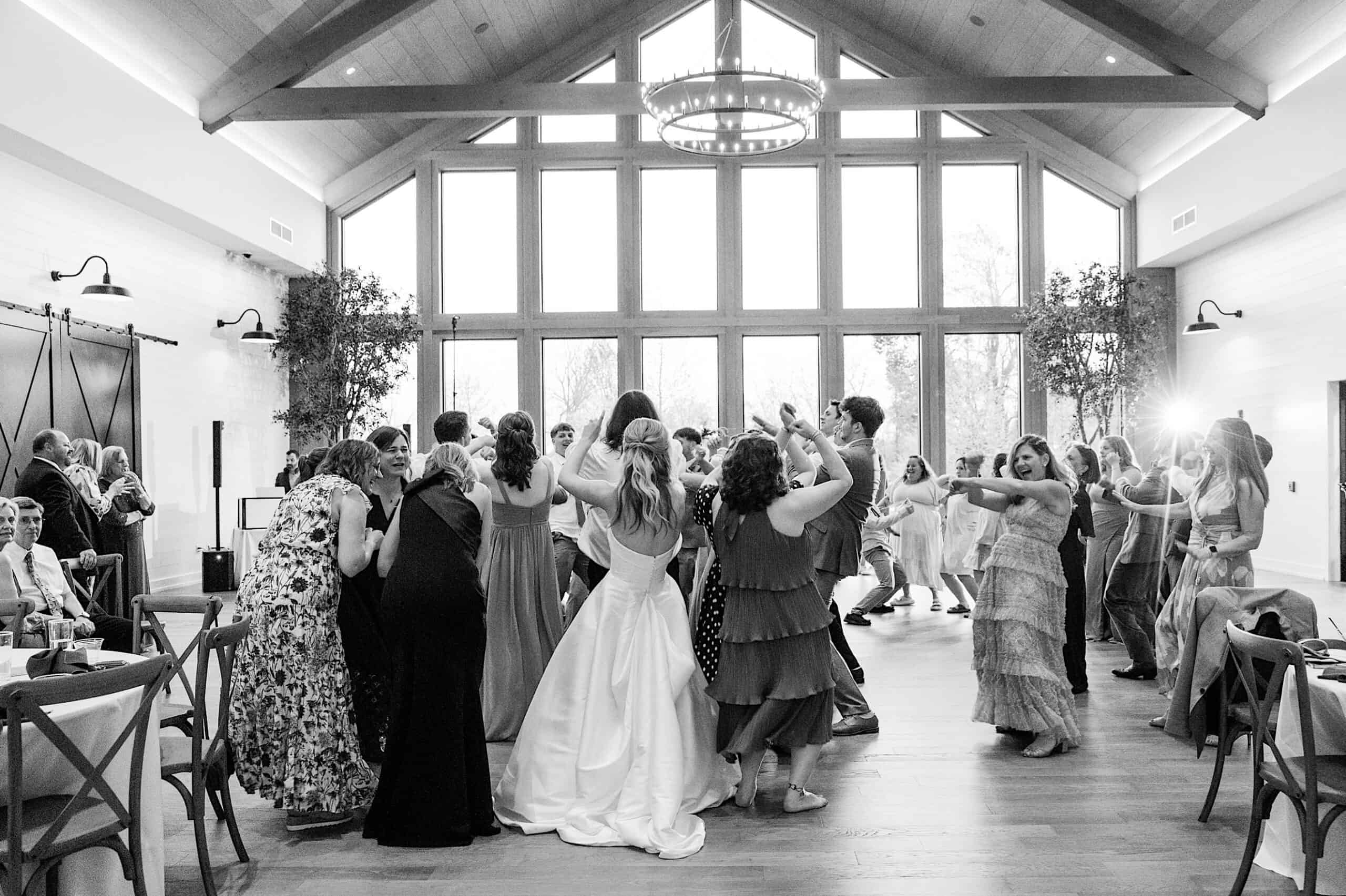 A group of people, including a bride in a white dress, dance together in a spacious, well-lit room with large windows and exposed beams at a Westwynd Gardens Spring Wedding.