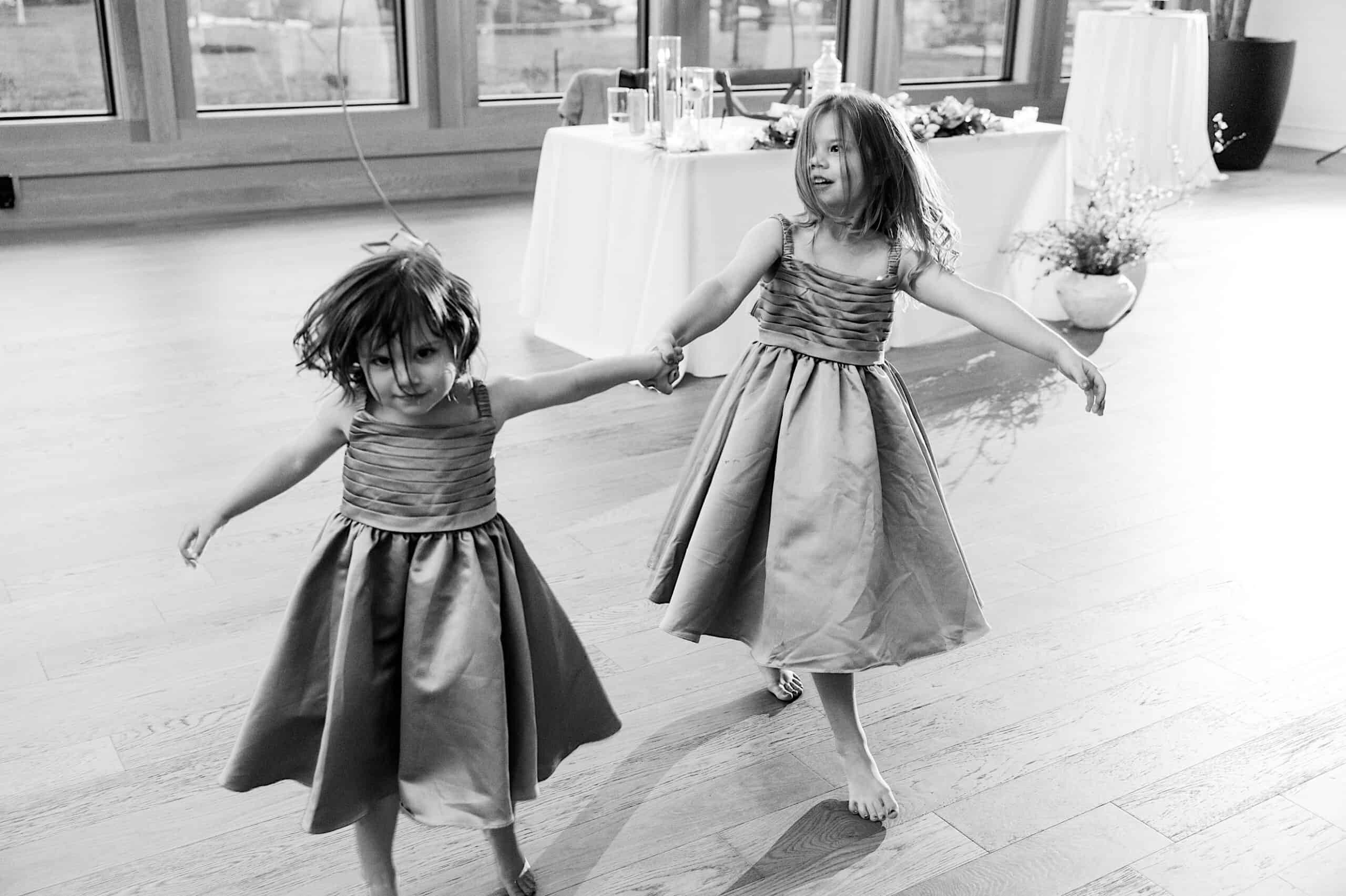Two young girls in matching dresses hold hands and spin barefoot on a wooden floor, sharing a joyful moment at a Westwynd Gardens Spring Wedding, with tables set in the bright room behind them.