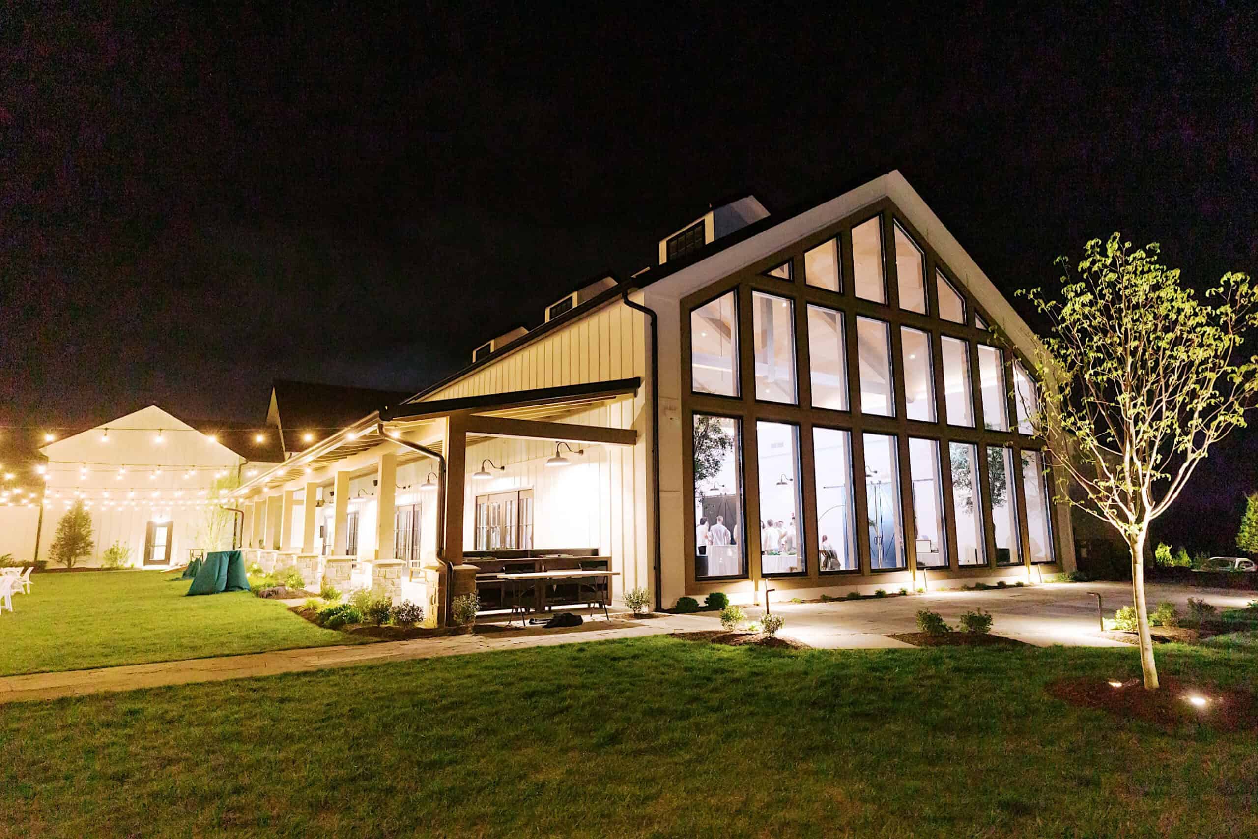 A modern barn-style event venue at Westwynd Gardens is lit up at night, with large windows, string lights, and a well-kept lawn—perfect for a dreamy Spring Wedding.
